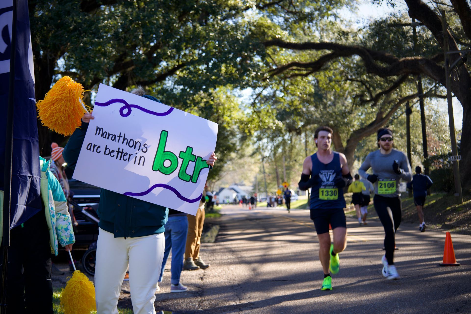 Marathon runners on a tree-lined road, with a volunteer holding a purple-and-white sign beside the course