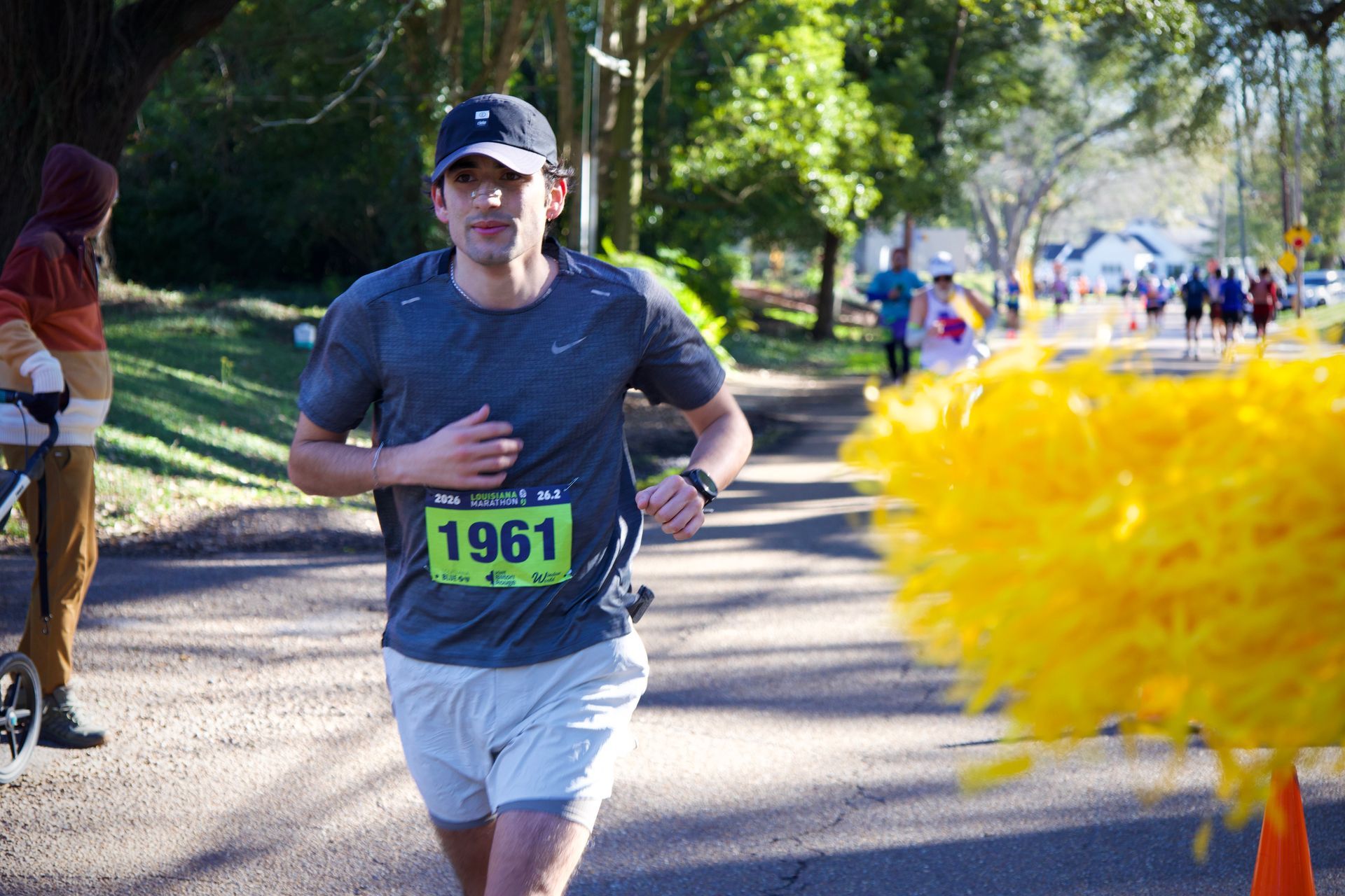 Runner in blue shirt and cap wearing bib 1961 on a sunny tree-lined road, with yellow pom-poms in foreground