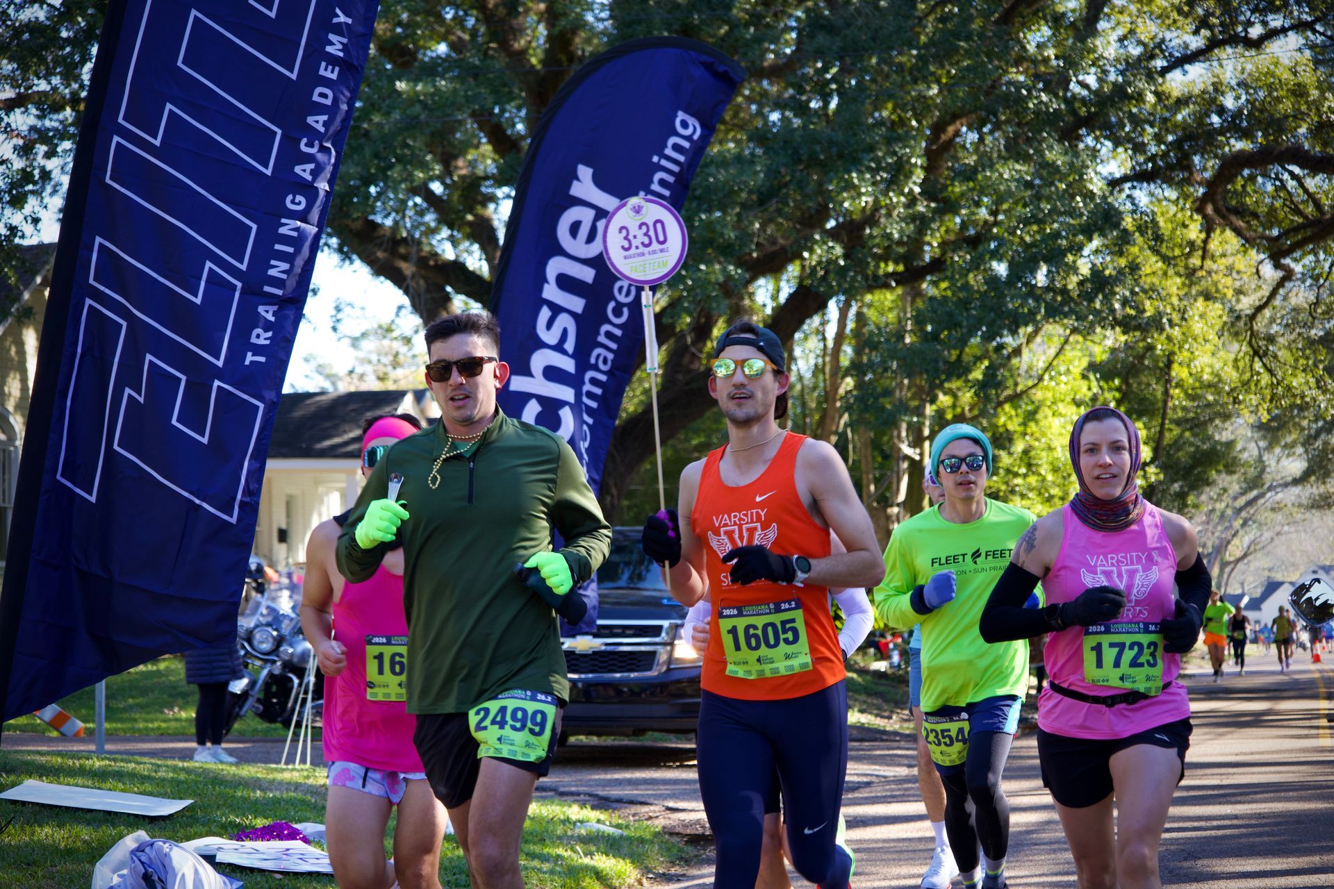 Runners in colorful shirts cross a finish line under a blue banner on a sunny outdoor path.