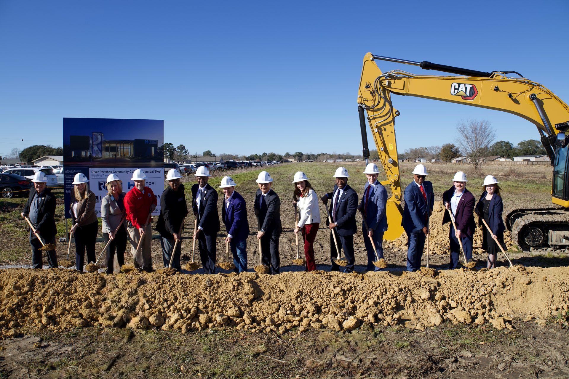 Group of people in hard hats standing beside an excavator at a groundbreaking site.