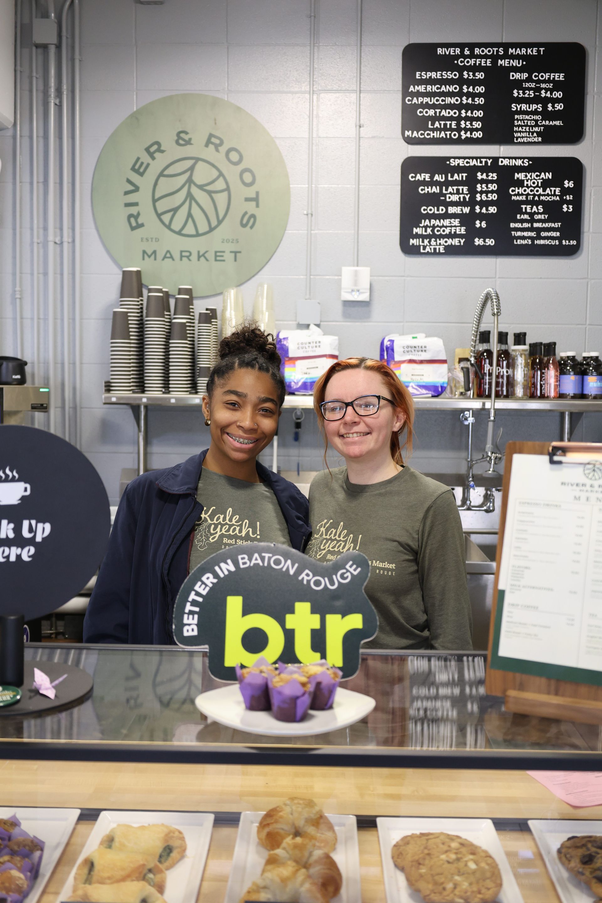 Two staff members smiling behind a bakery counter with purple pastries and a round logo sign.