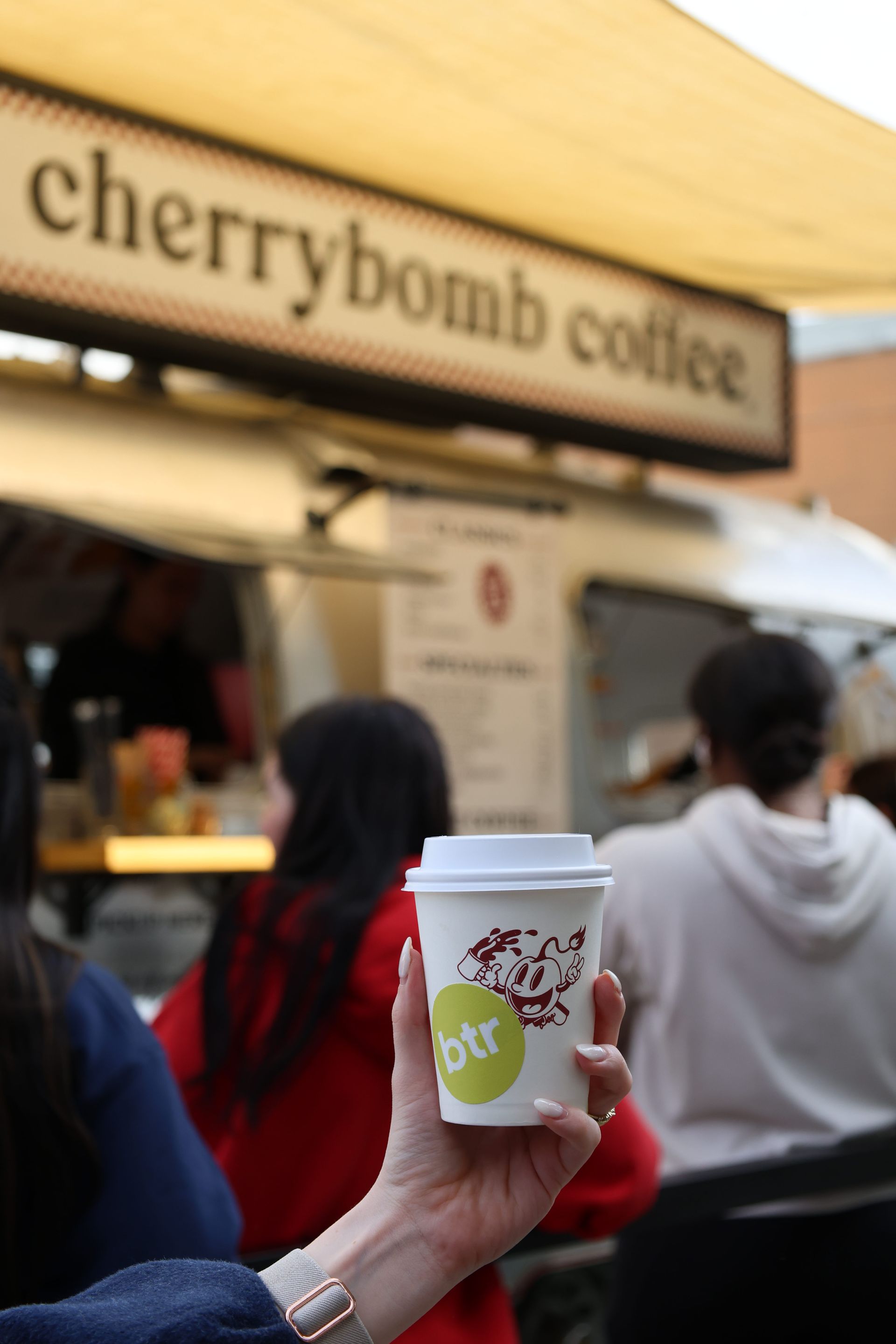 Hand holding a Cherrybomb Coffee cup at an outdoor café counter, with people in line behind it