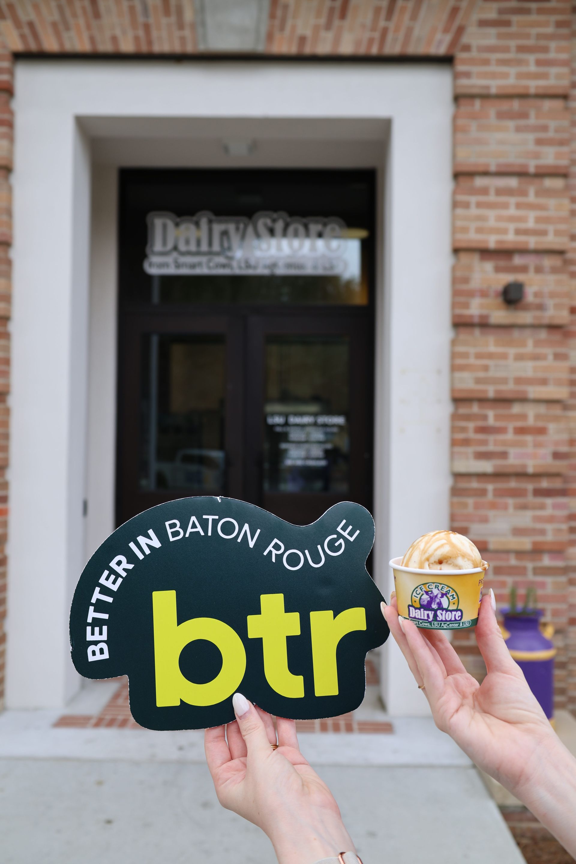 Hands holding Better in Baton Rouge sign and dessert in front of a Daily Stop storefront
