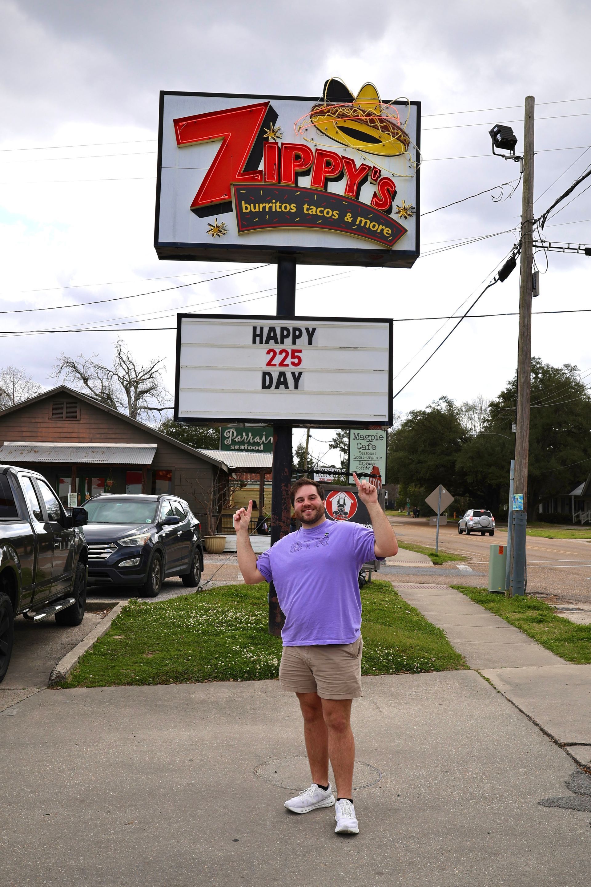 Person posing under a Zipps bar sign on a sidewalk, wearing a purple shirt and khaki shorts.