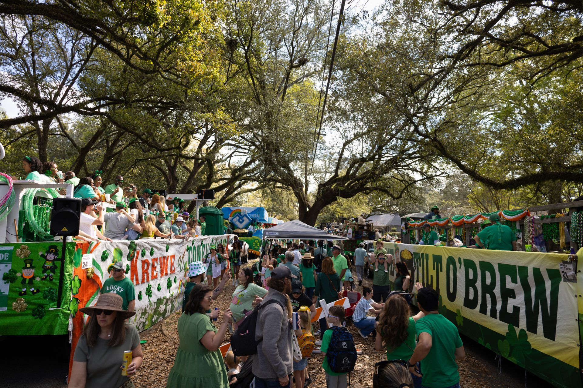 Crowd at a green-themed outdoor festival under trees, with banners and people walking among booths