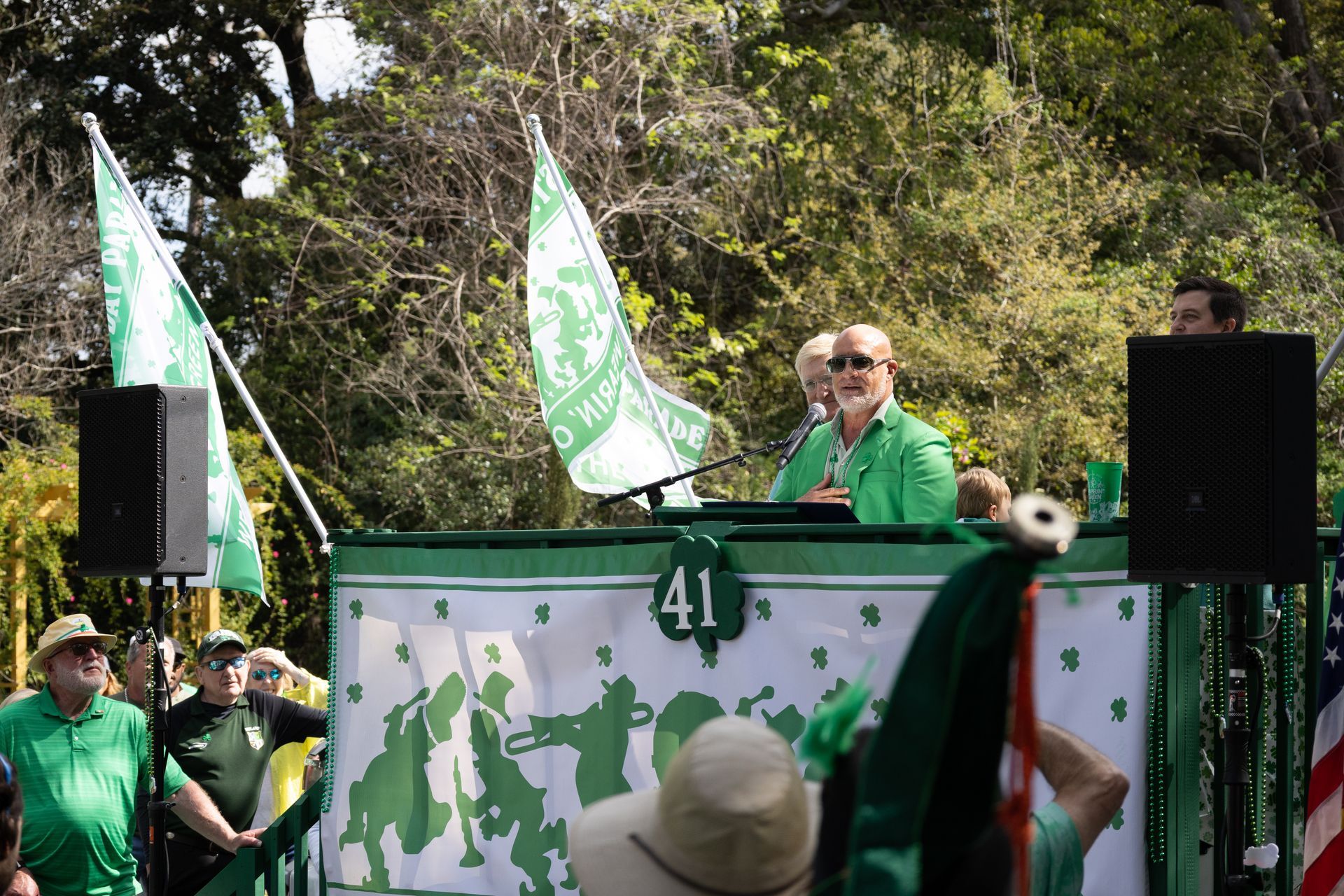 Speaker in green at podium with flags and crowd outdoors