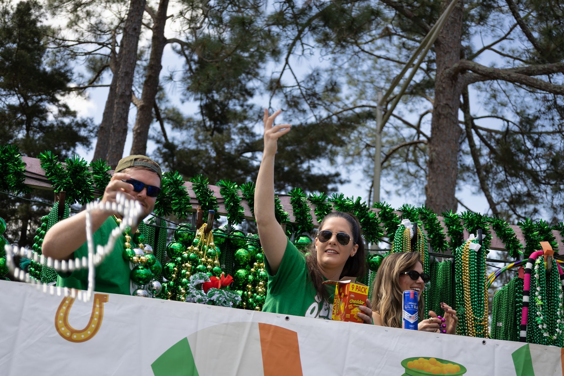 Two people in green shirts and sunglasses waving at a parade float decorated with colorful beads outdoors