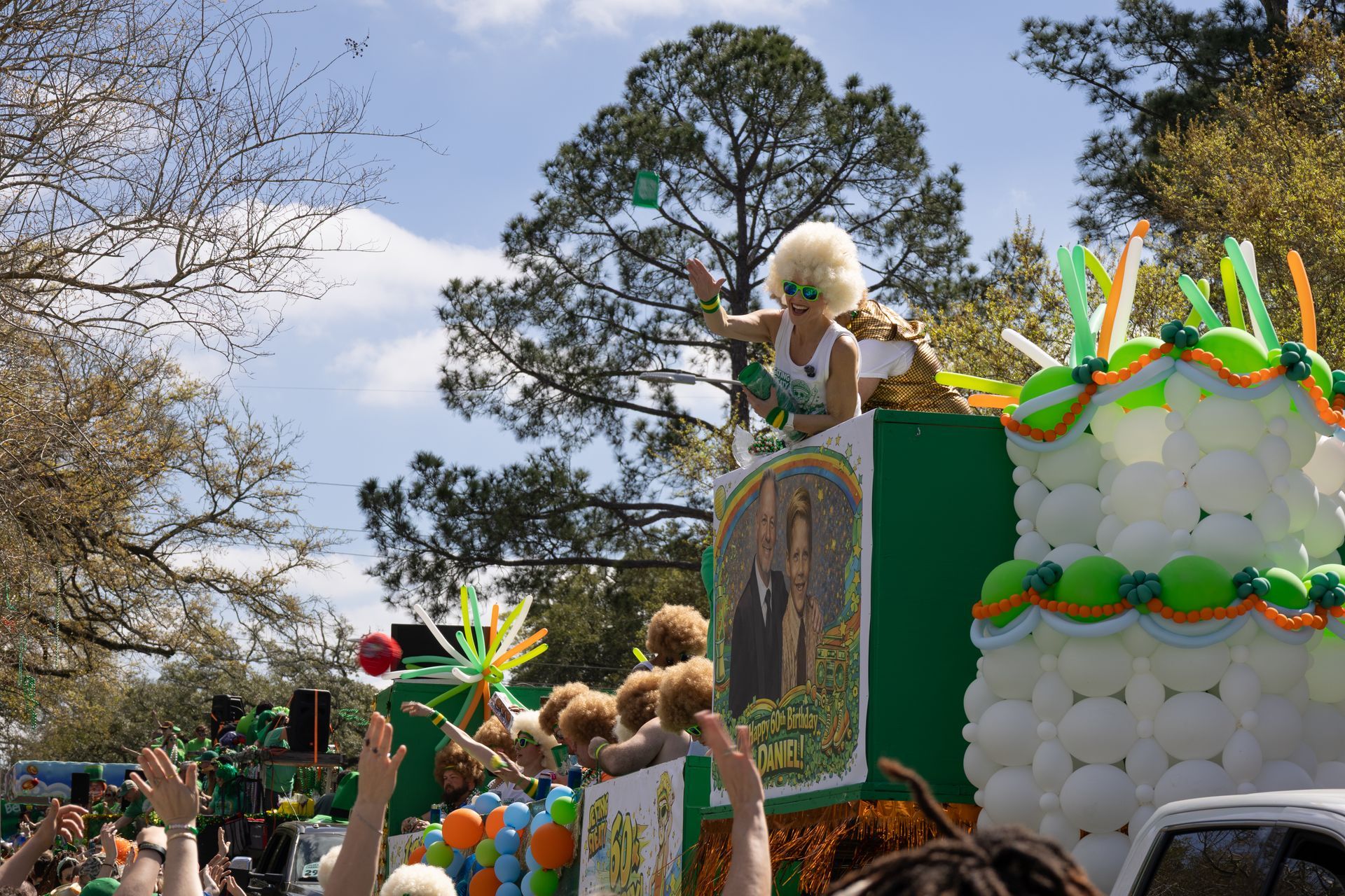 Colorful parade float with teddy bears, balloons, and green and orange decorations under trees and sky