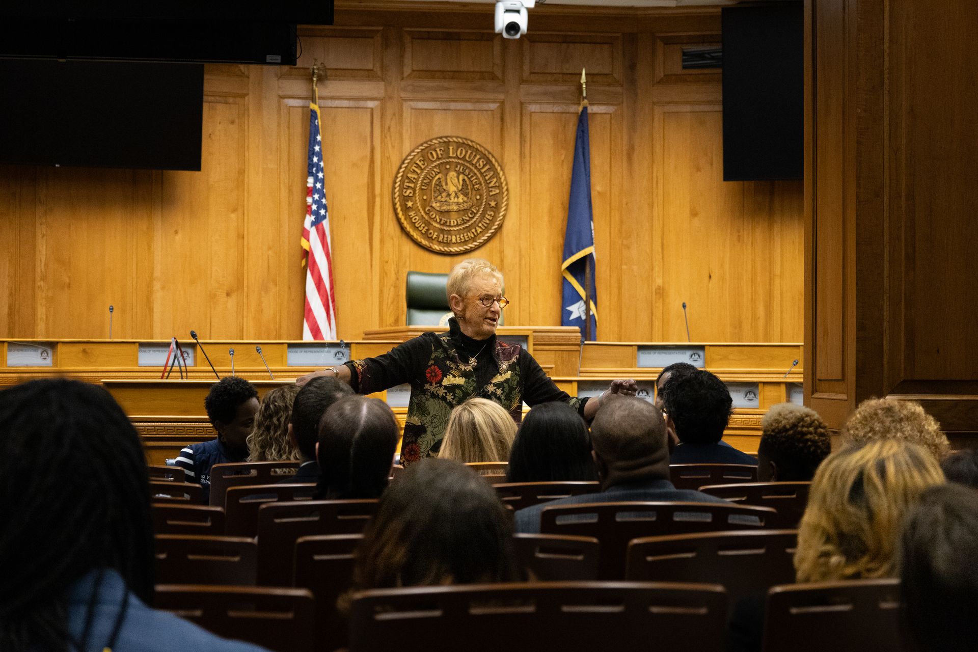 Speaker addressing an audience in a wood-paneled courtroom with U.S. and state flags behind them