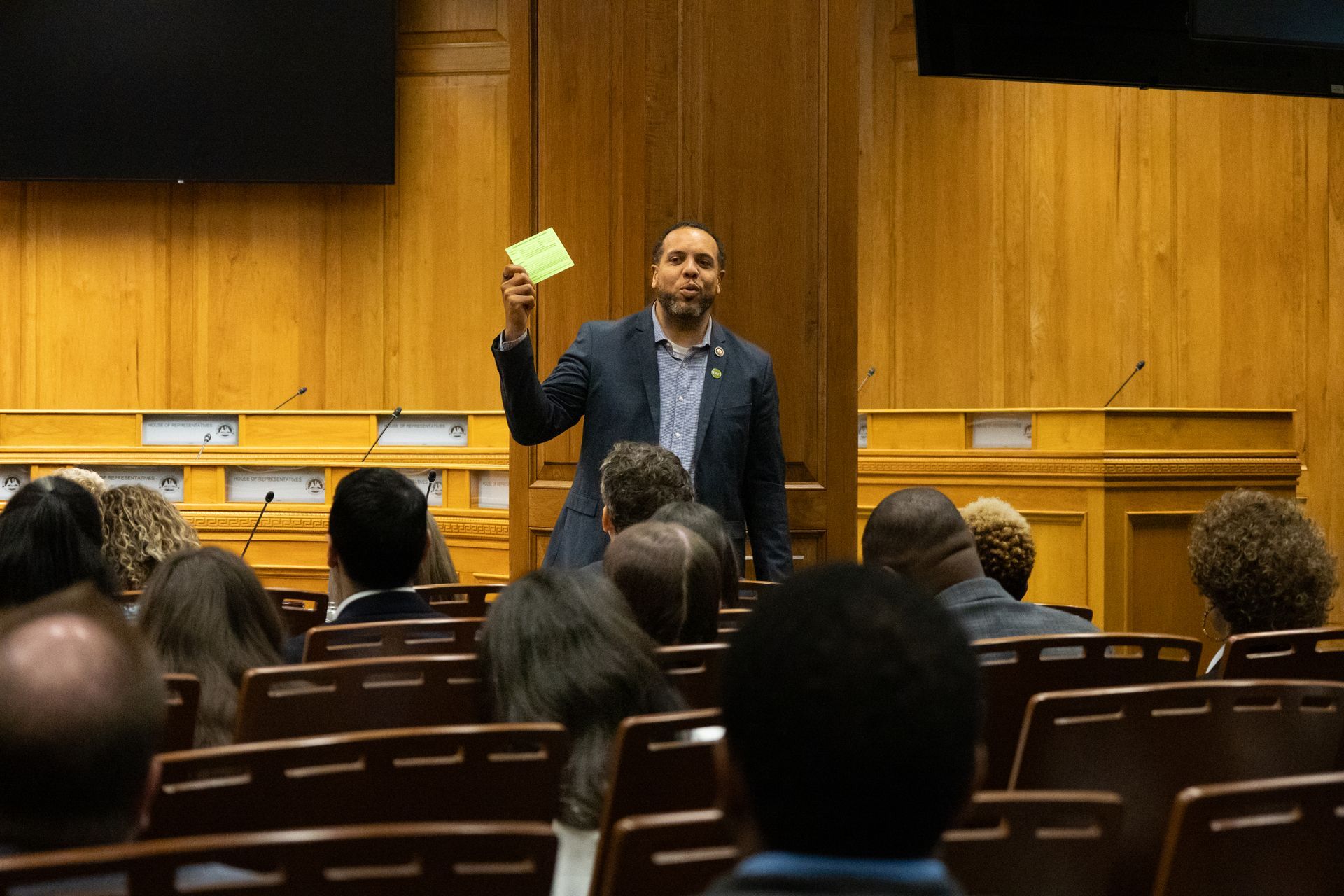 Speaker holding a note card addresses an audience in a wood-paneled lecture hall.