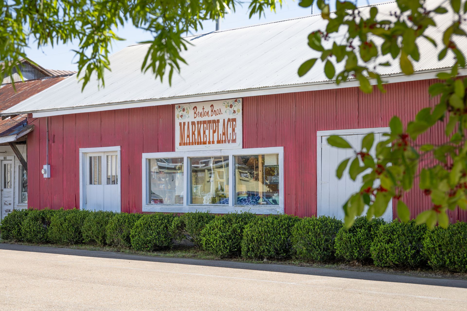 Red storefront building with white trim, large windows, and bushes along the front under leafy branches.