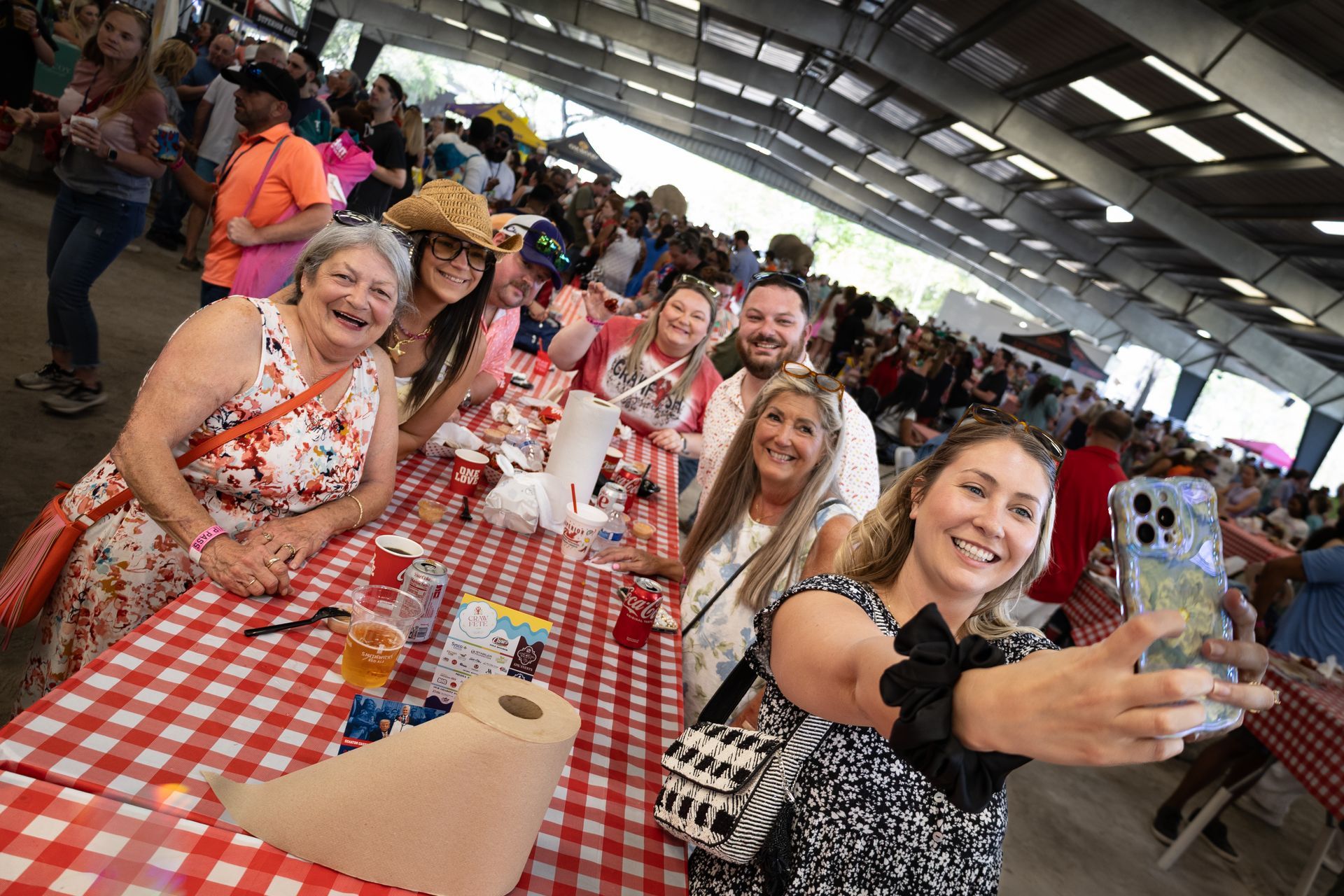 Group of people at a long picnic table in a crowded indoor hall, smiling and taking a selfie at a red-checkered tablecloth