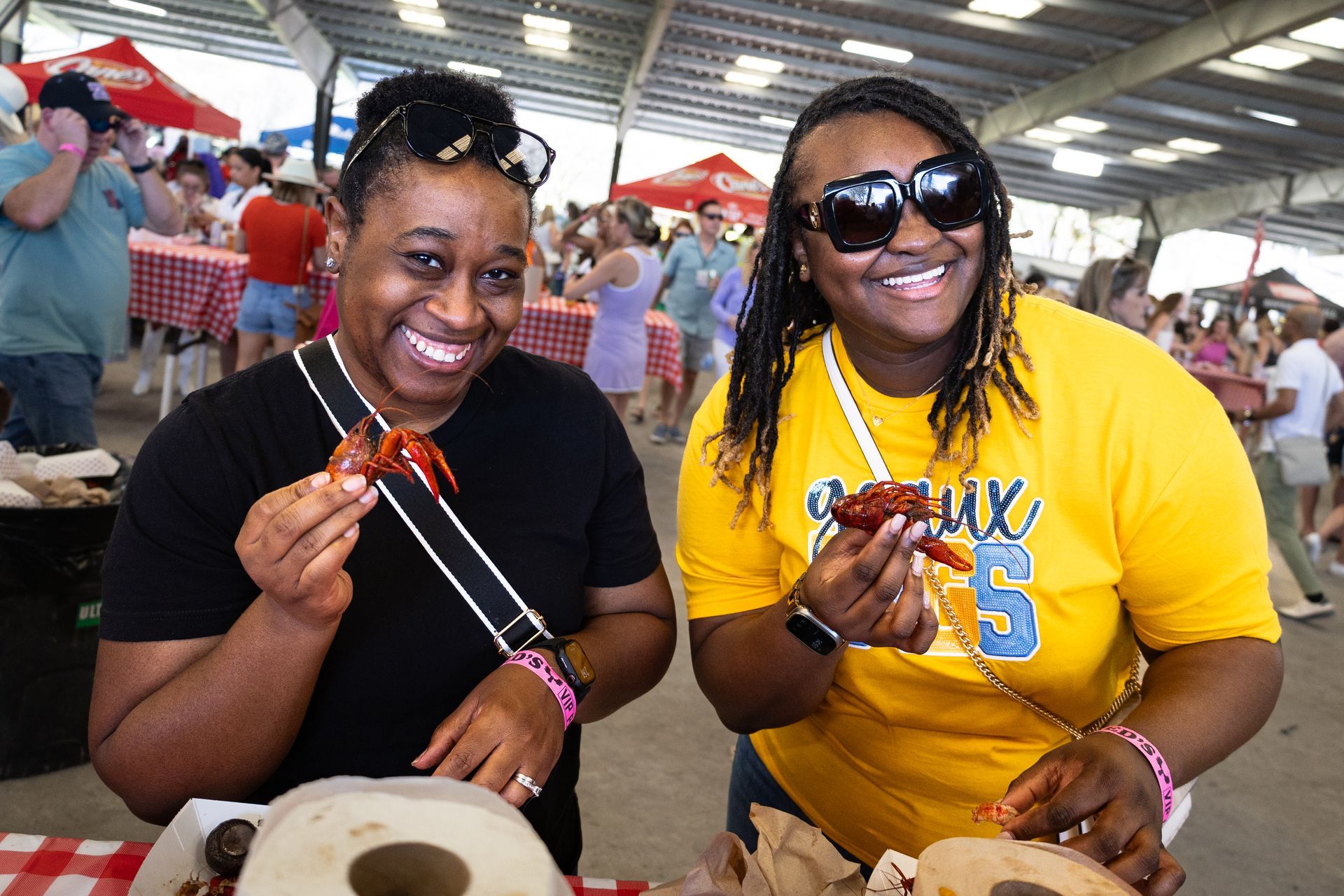 Two women smiling and eating crab legs at an indoor seafood market, one in yellow and one in black.