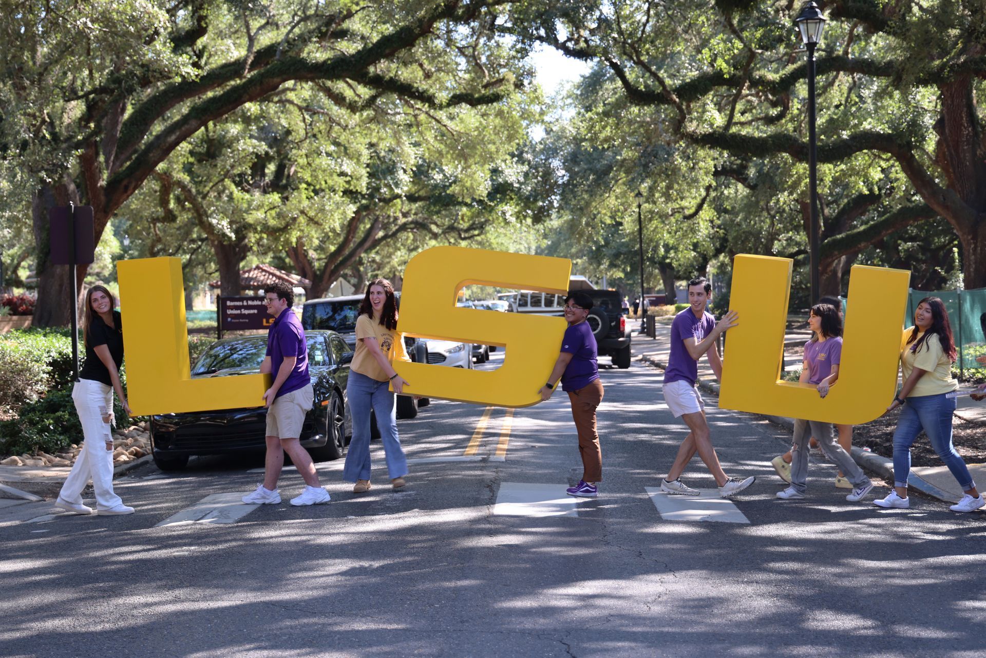 People carrying large yellow “LSU” letters during a campus parade under trees.