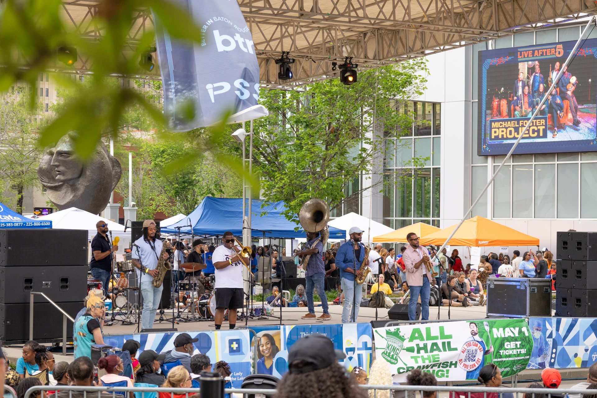 Outdoor band performing on a stage at a lively crowd event with a large screen and trees nearby