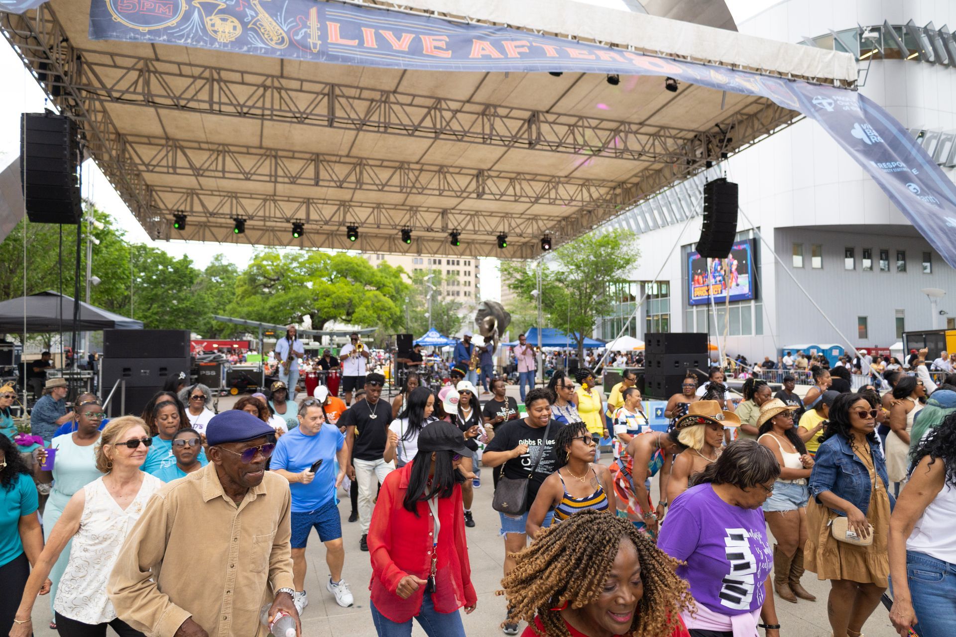 Crowd walking under a large outdoor stage canopy at a public event, with people in colorful clothing.