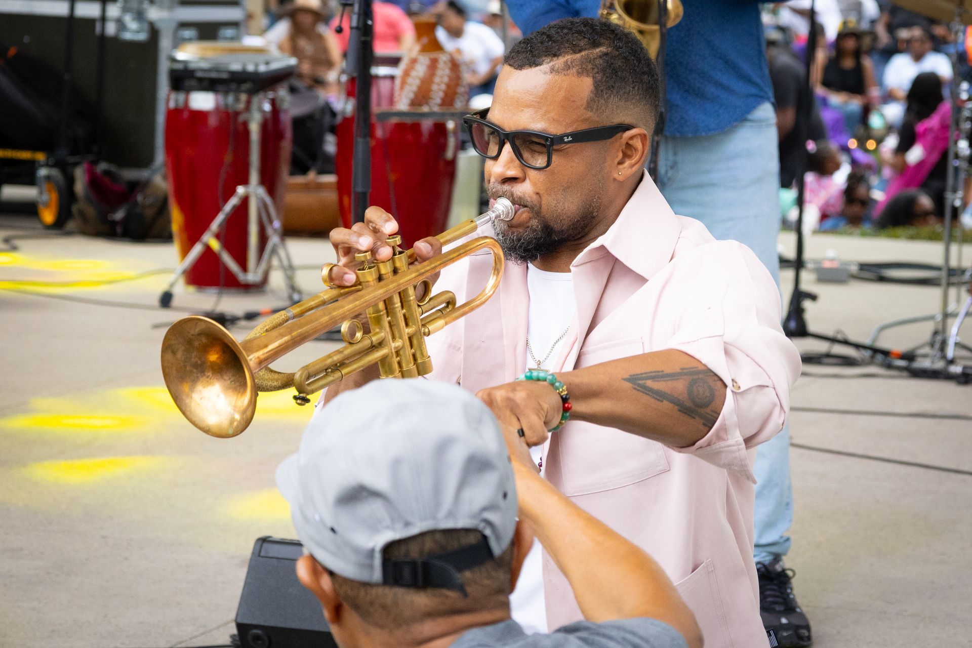 Trumpeter playing at an outdoor festival, wearing glasses and a pale pink shirt, with drums in the background