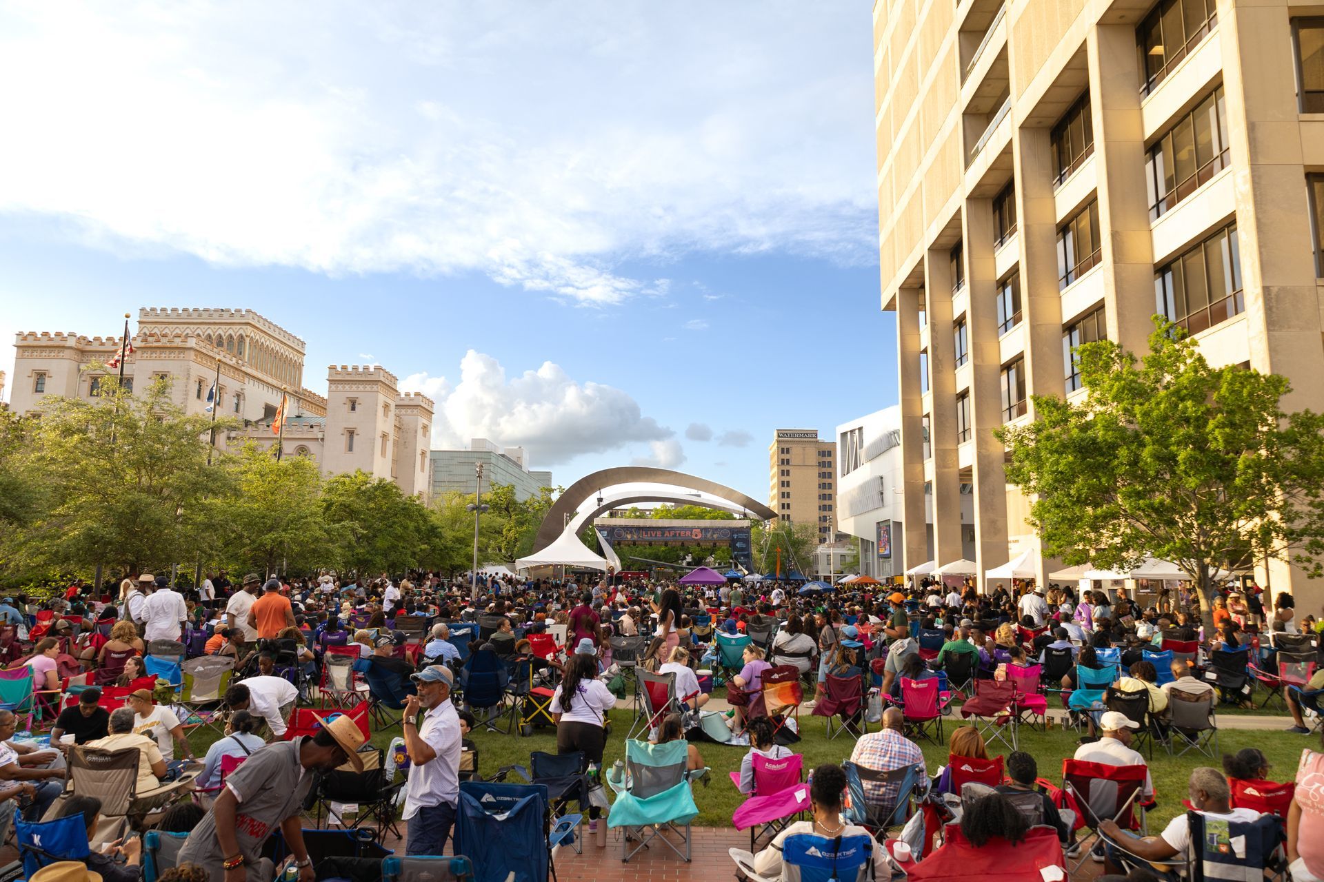 Crowded outdoor concert in a city plaza with seated people, trees, and tall buildings under a blue sky