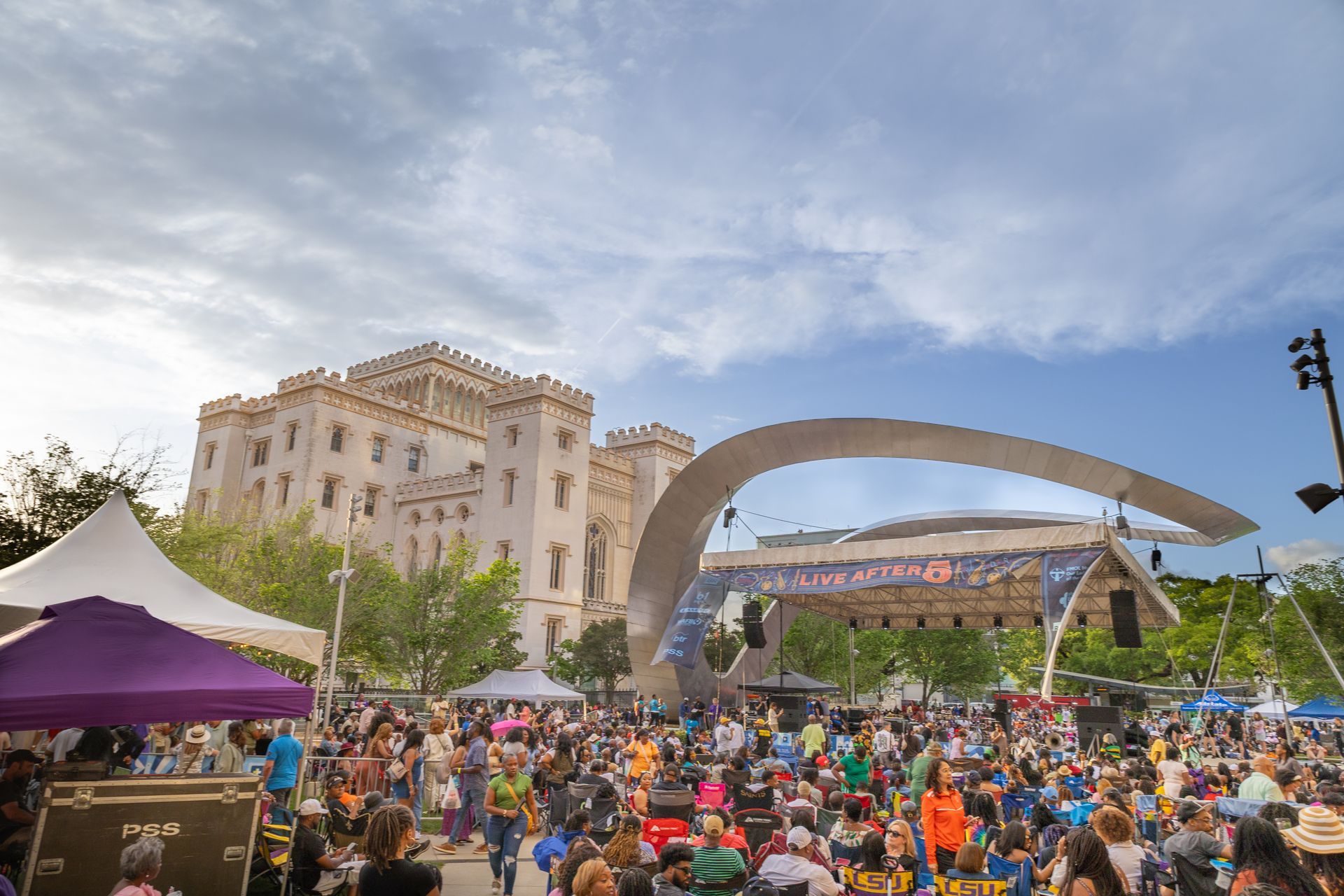 Crowd gathered at an outdoor concert with a stage, white tent, and tall buildings under a blue sky.