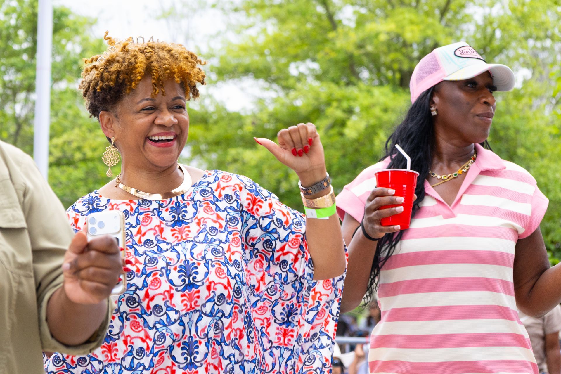 Two smiling people outdoors holding drinks, one dancing in a blue floral shirt and the other in a pink striped shirt.