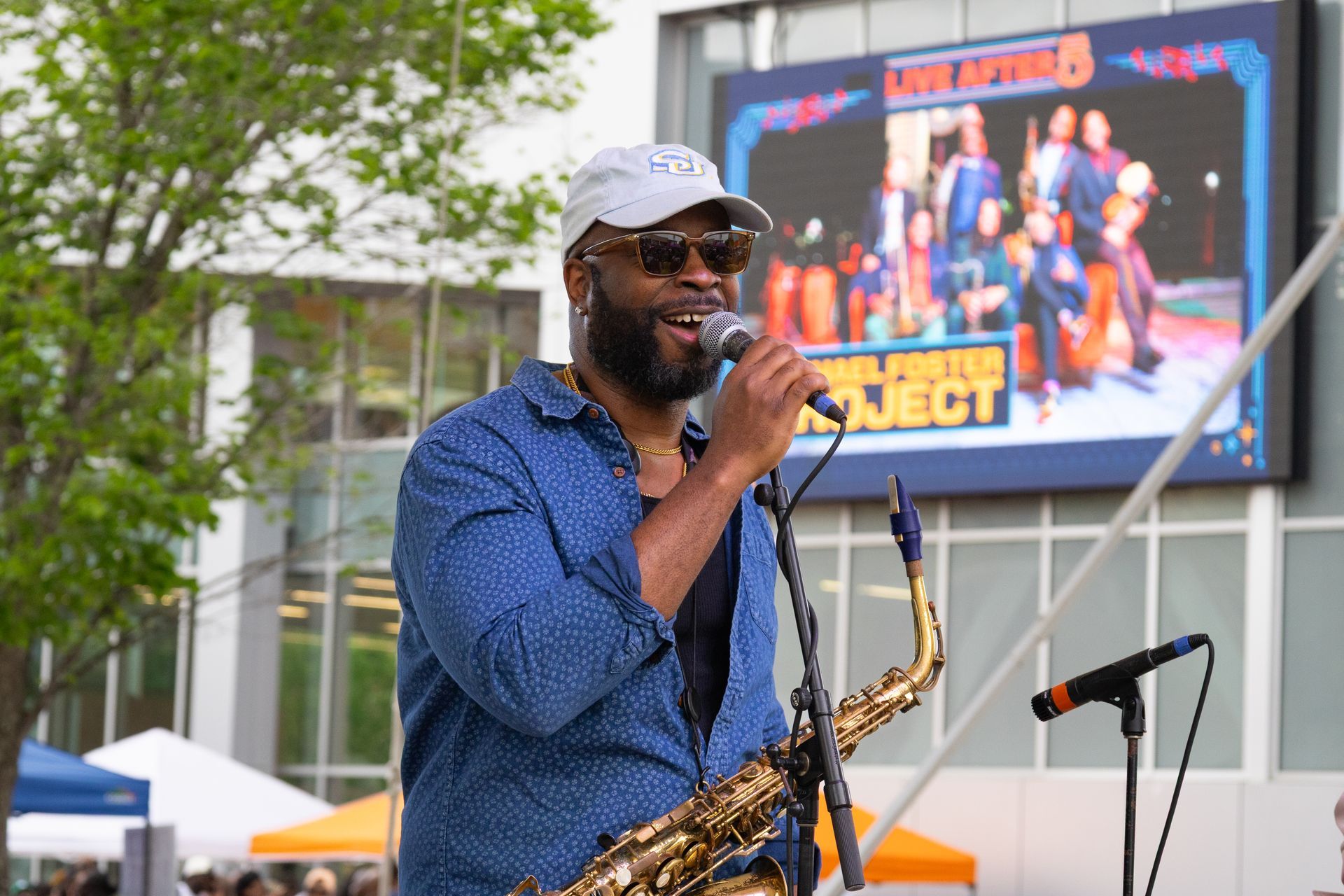 Man singing into a microphone with a saxophone at an outdoor stage event