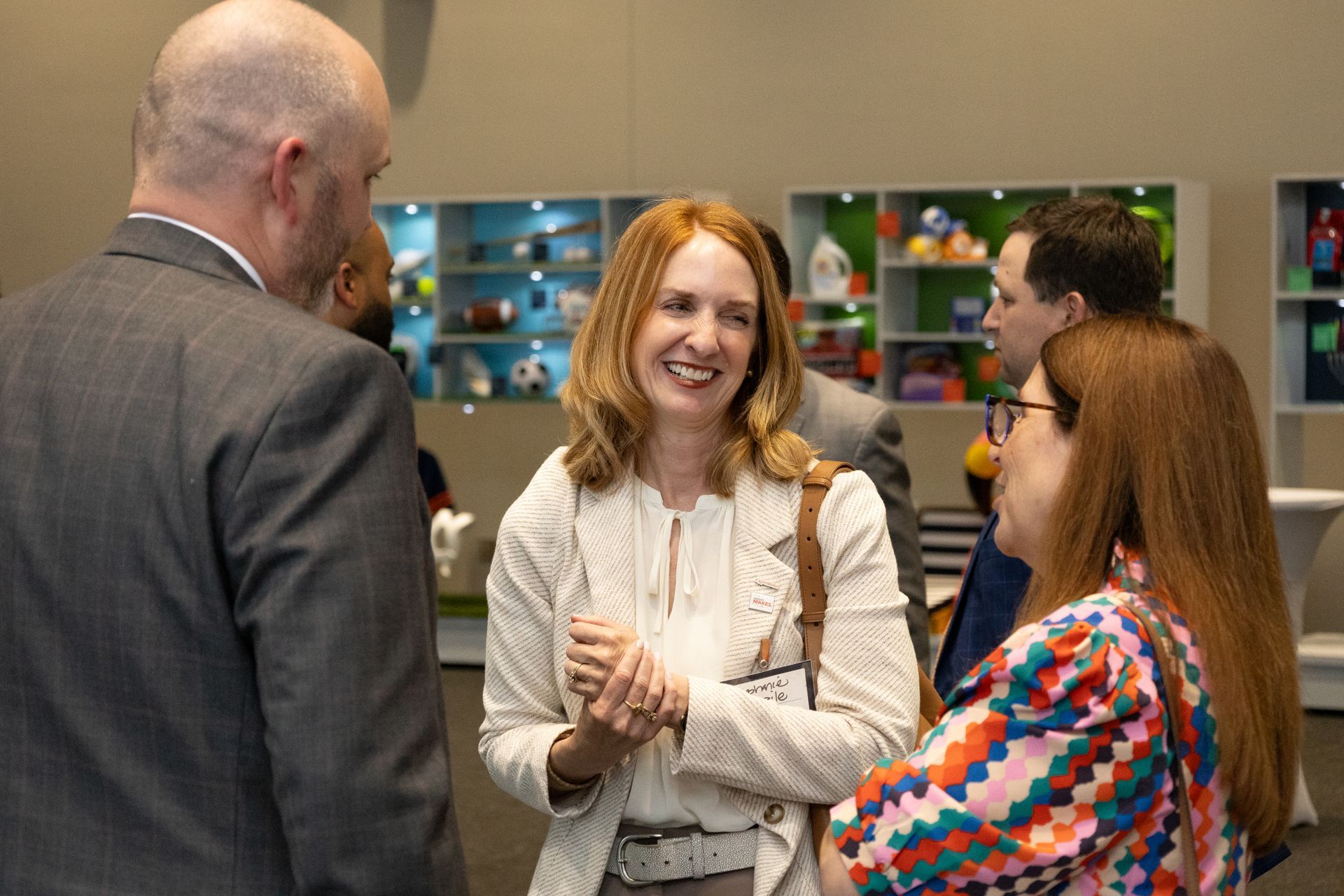Three people chatting and smiling at an indoor event, with one man in a suit and two women in the foreground.