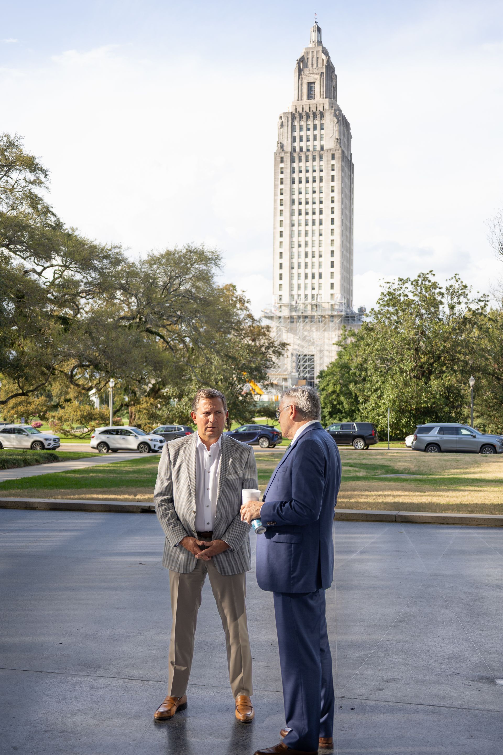 Two men in suits talk on a sidewalk with a tall historic tower and trees behind them.