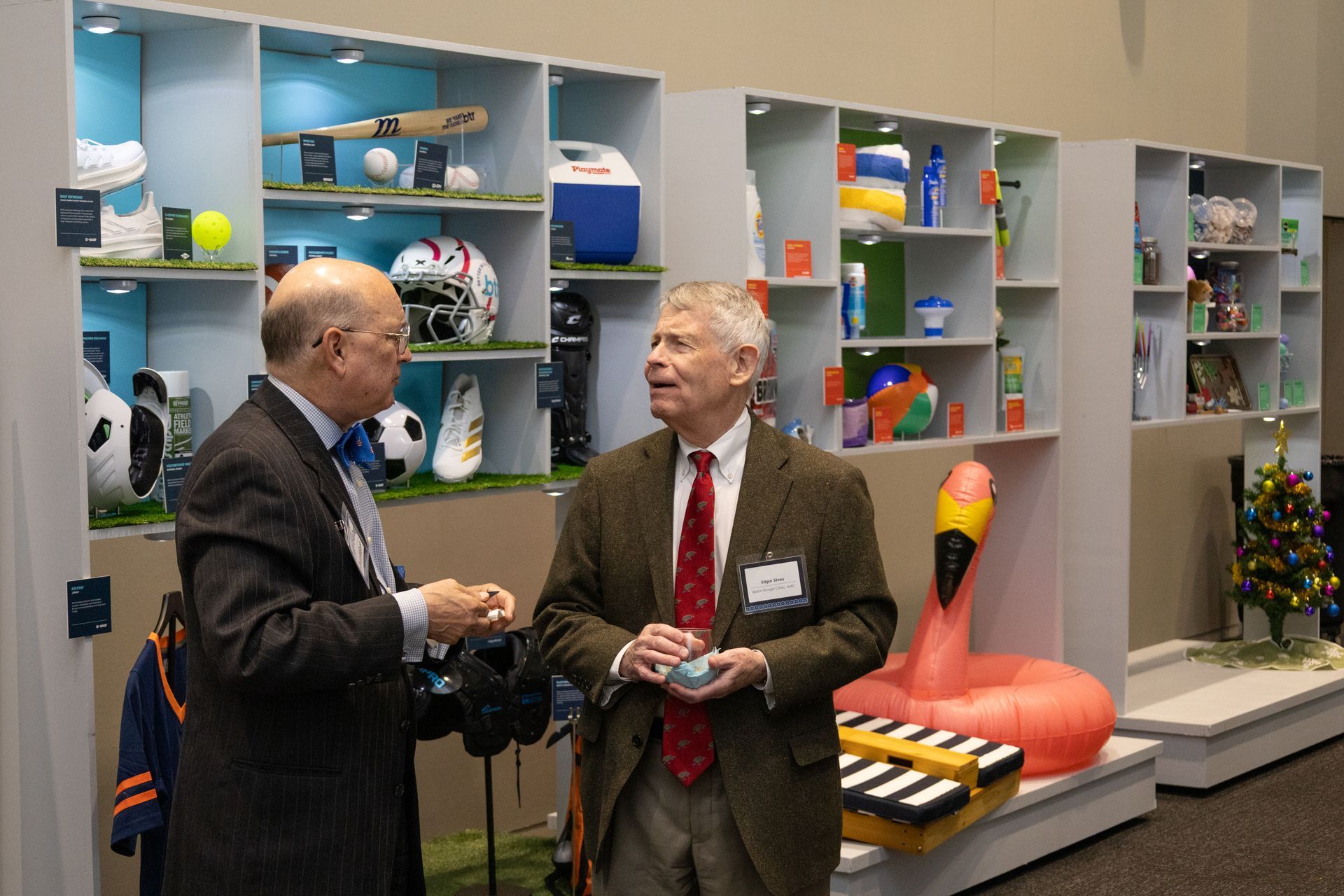 Two men talking in a colorful office display area with shelves, toys, and a large orange chair.
