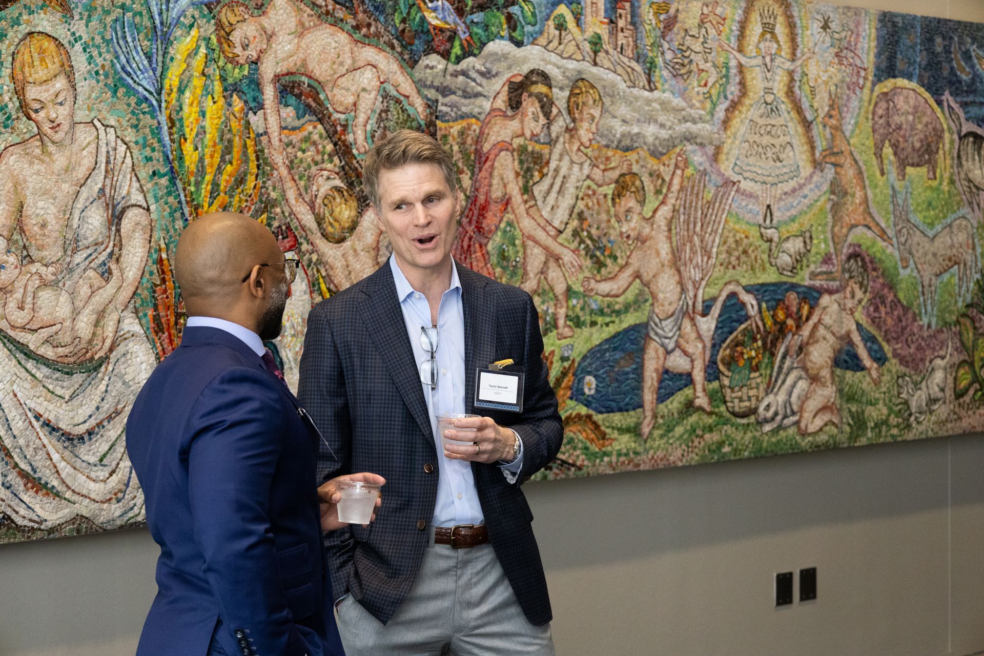 Two people converse in a gallery beside a colorful mural and a wall-mounted display.