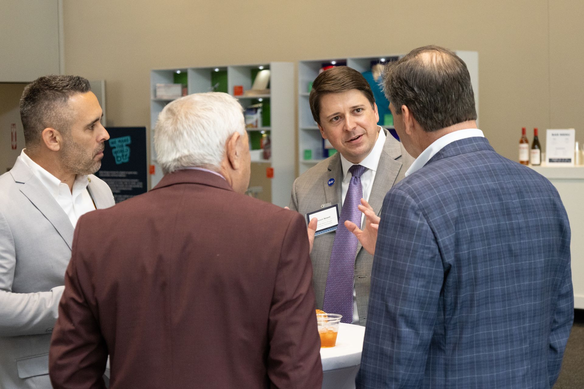 Four people in business attire talk in an office hallway, gathered around a table and papers.