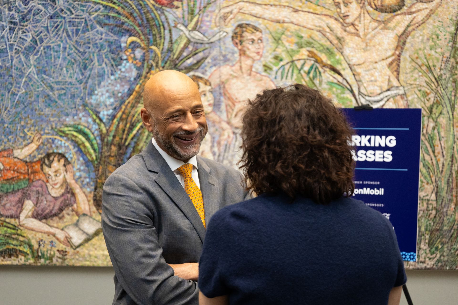 Two people shaking hands in front of a colorful mural and a sign reading “Working Progress.”