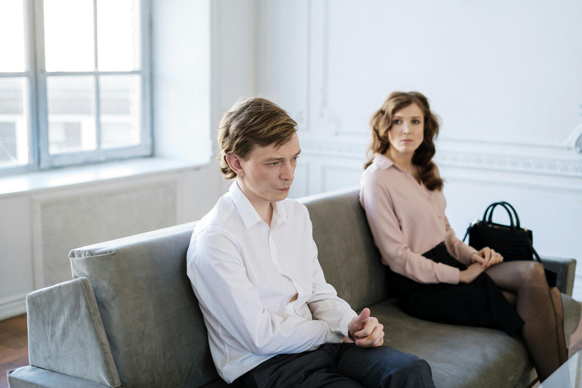 Two people sitting on a couch in a bright room, one looking down and the other facing forward