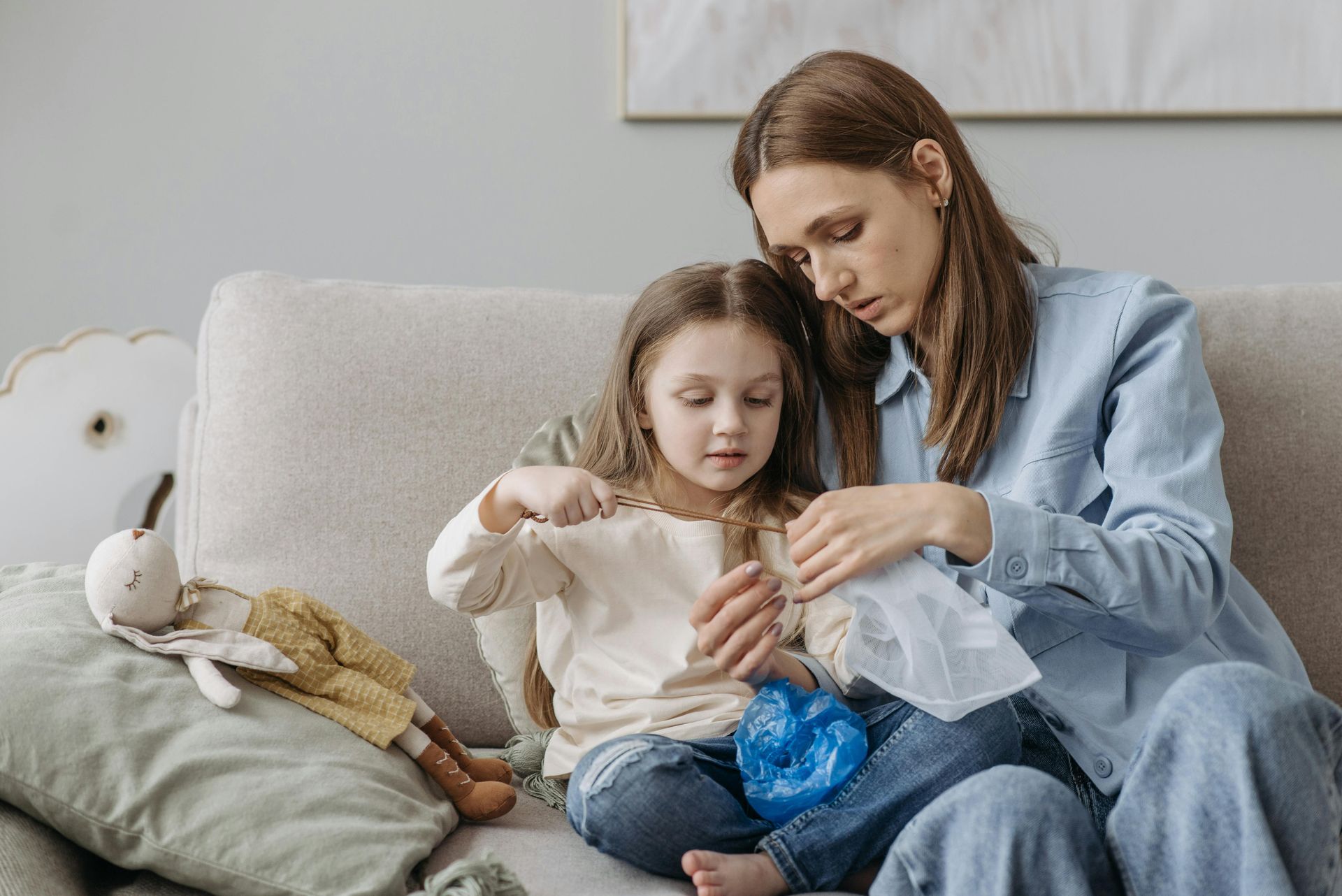 Woman and child making a blue craft project on a light gray sofa, with stuffed toys nearby.