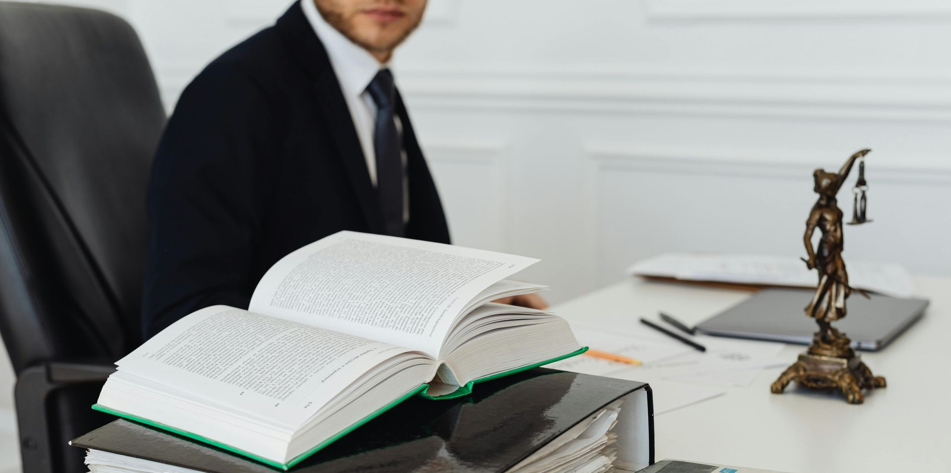 Open book on desk in an office, with a man in a suit and a small justice statue nearby