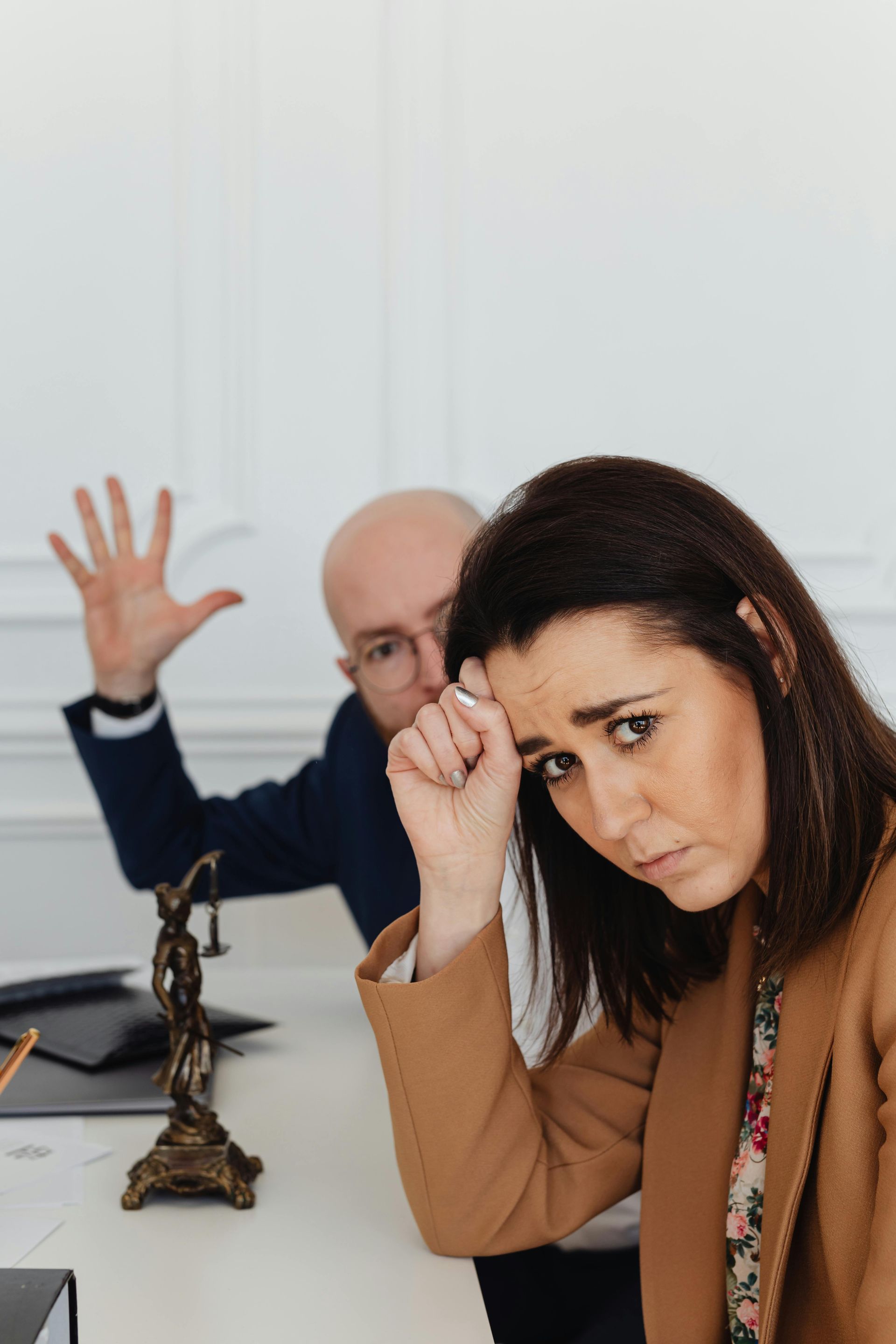 Woman in foreground with hand to forehead, looking worried, while man in background raises his hand in an office.
