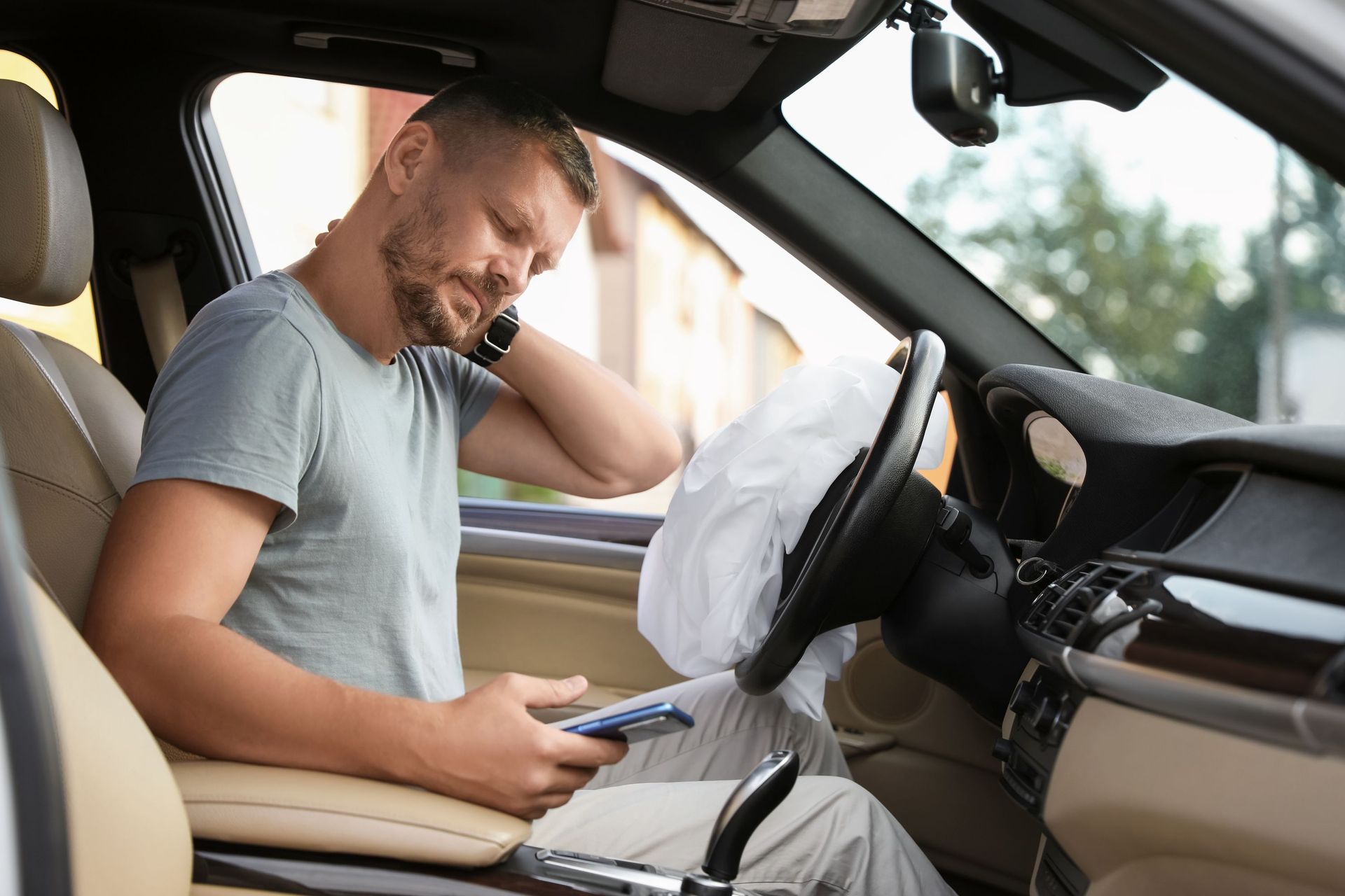Man in a car holding a phone, looking uncomfortable and clutching his neck, with a deployed airbag visible.