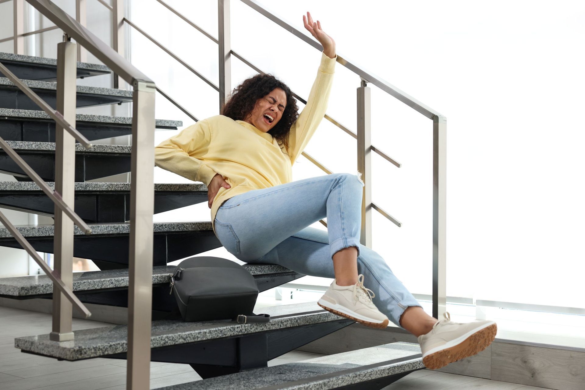 Woman in a yellow sweater slides down indoor stairs, smiling and raising one arm.