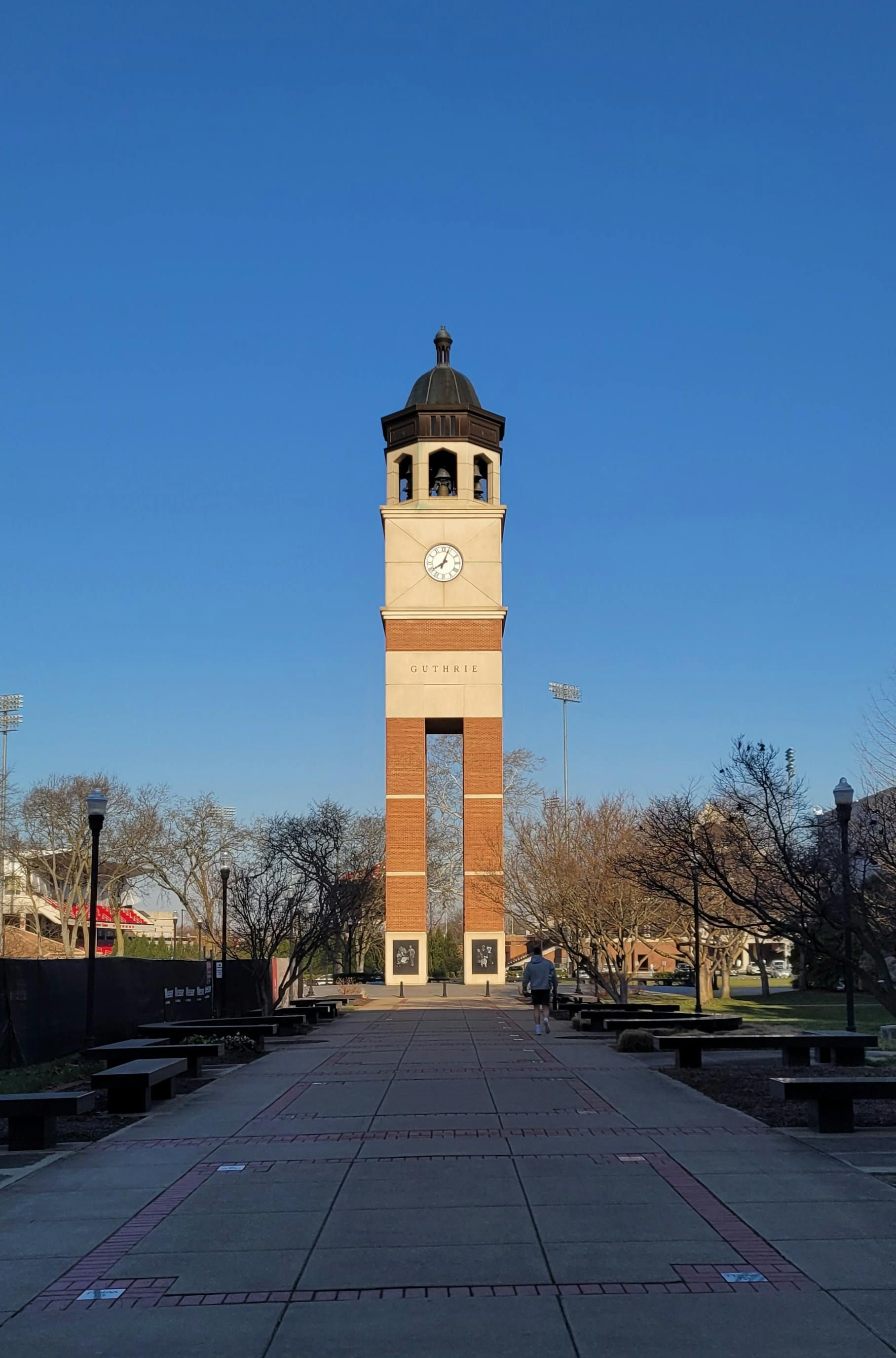 Clock tower of brick and beige, with clock face, against blue sky. Walkway lined with benches.