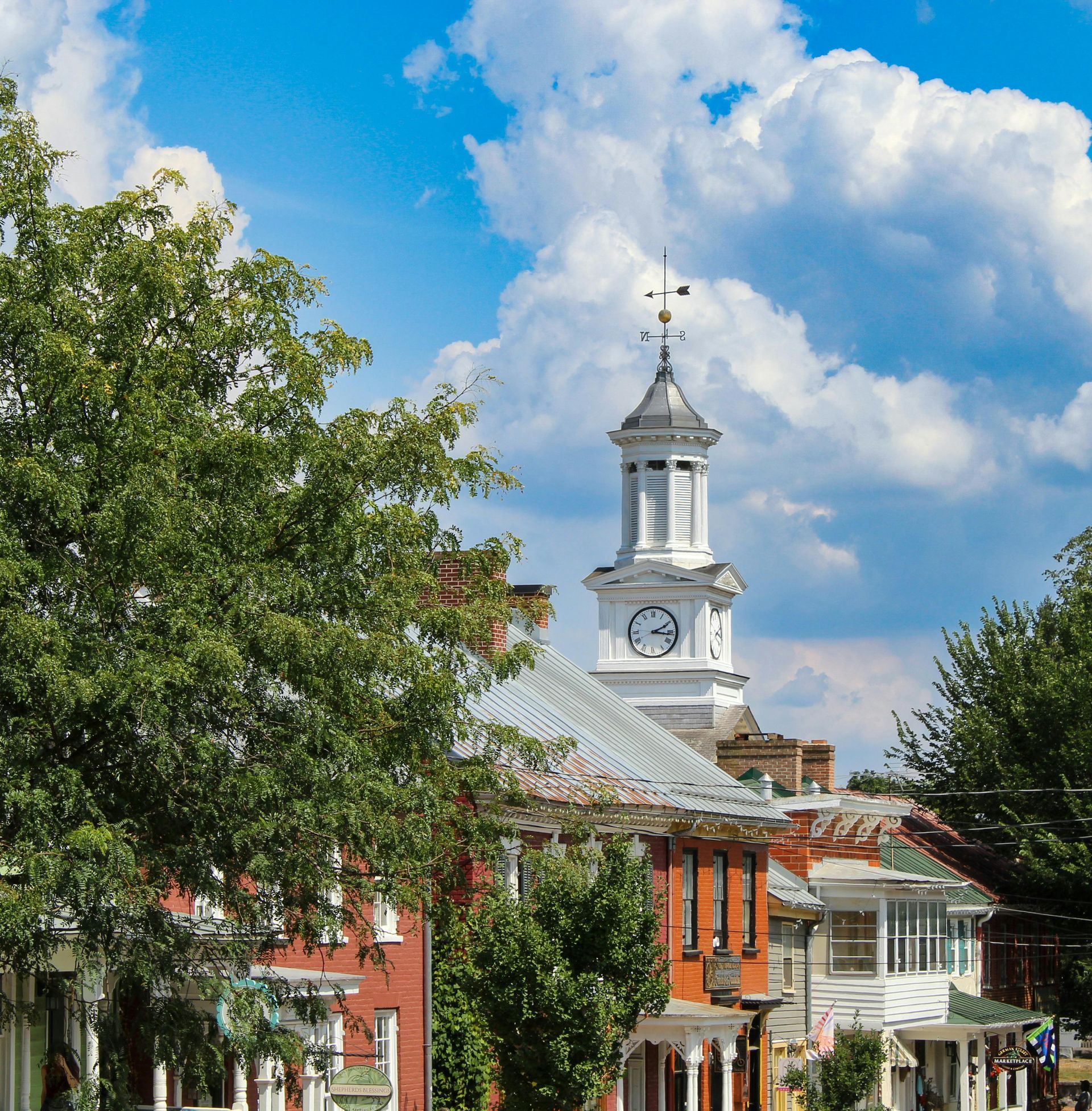 Clock tower atop a building in a historic town, blue sky and clouds, green trees.