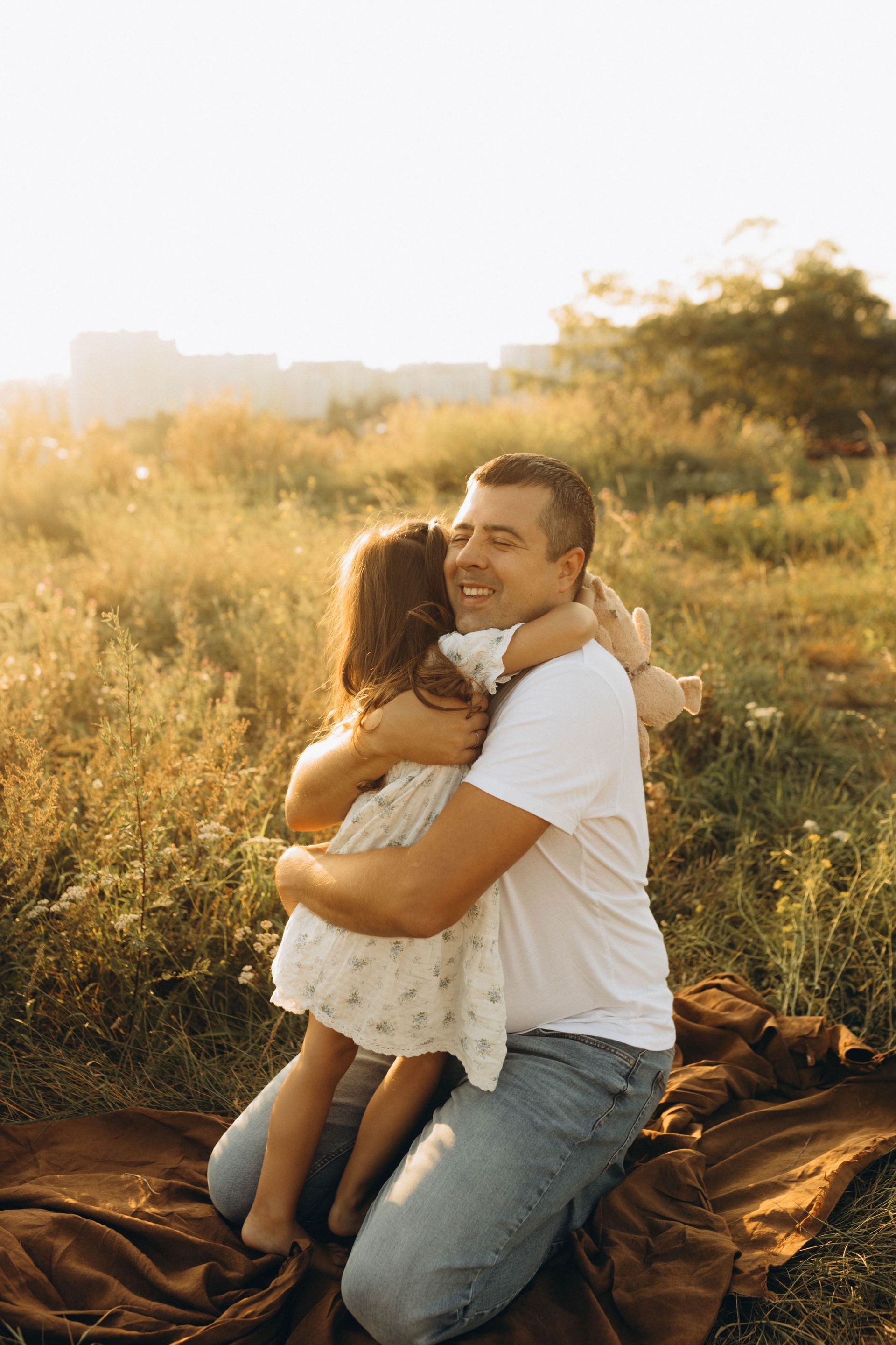 Man kneels in a sunlit field hugging a child in a white dress