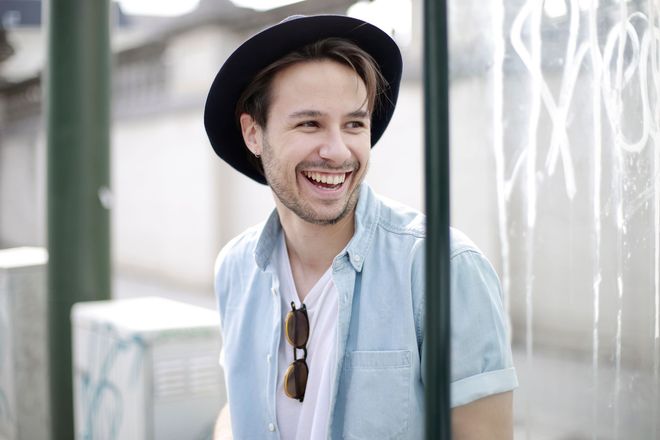 Smiling person in a black hat and light blue shirt standing behind metal bars outdoors