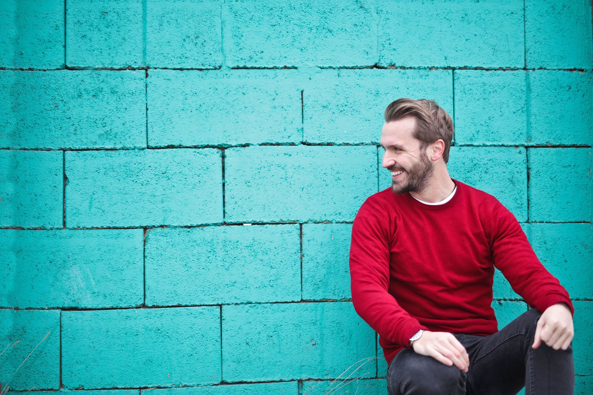 Man in a red sweater sitting against a turquoise brick wall, looking to the side with a slight smile
