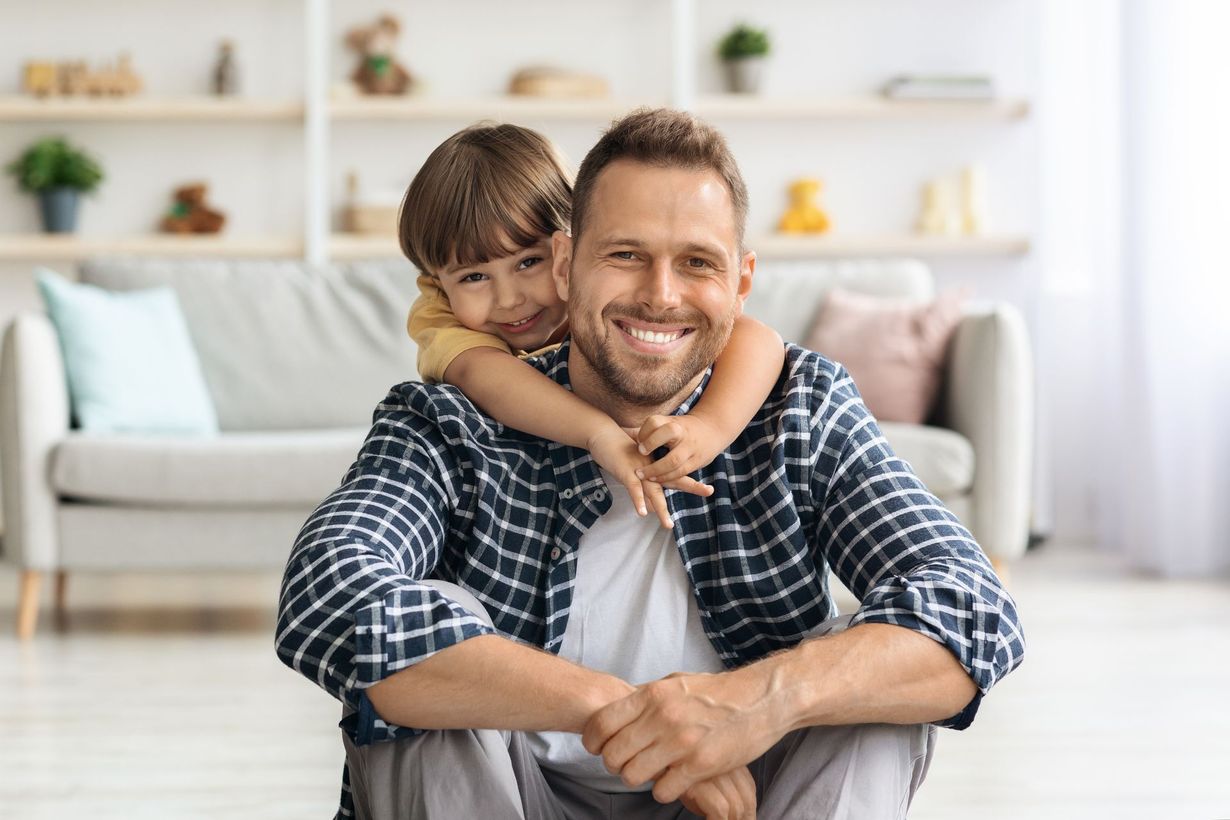 Smiling adult with a child on his back in a bright living room