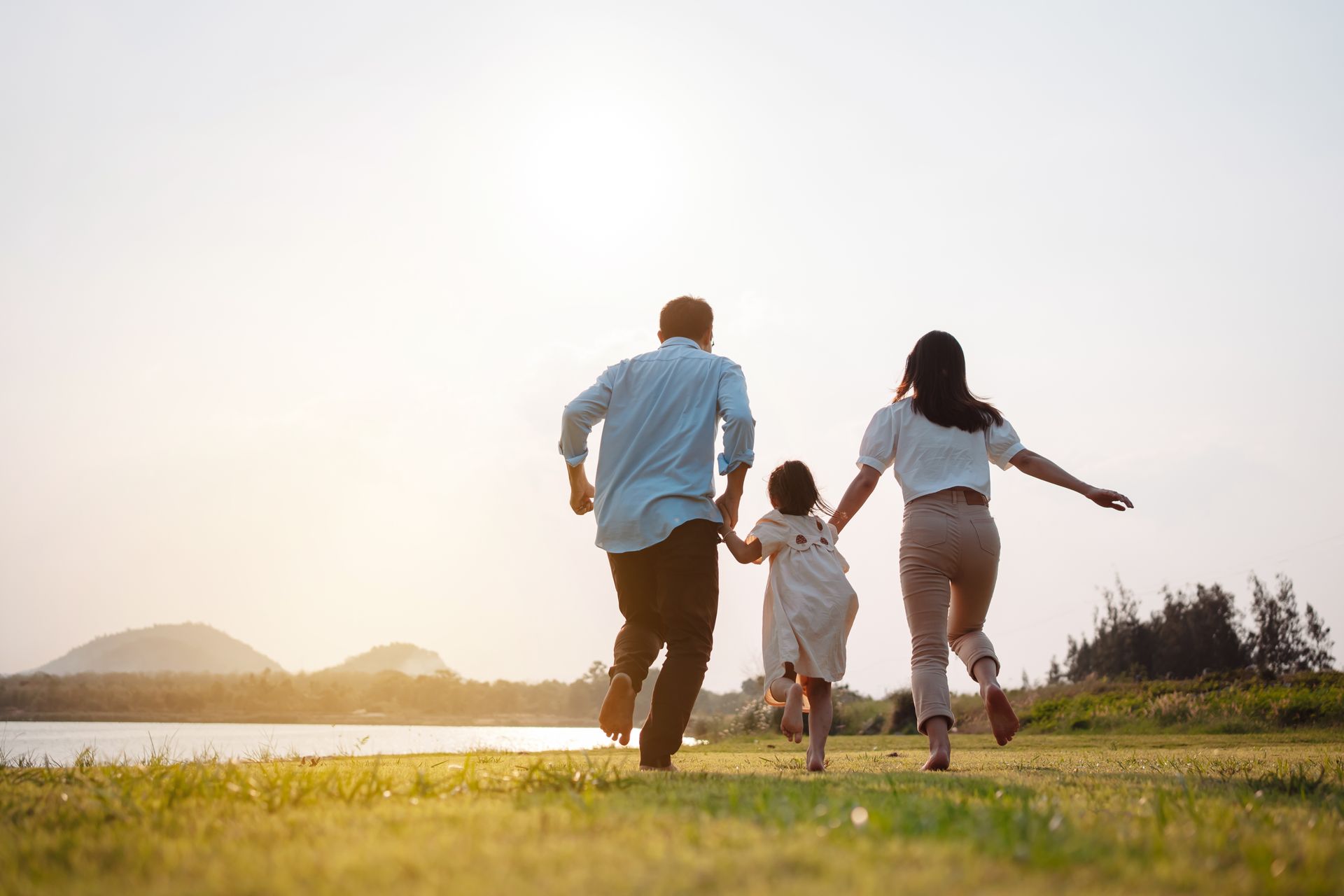 Three people running hand in hand through a sunny grassy field near water at sunset