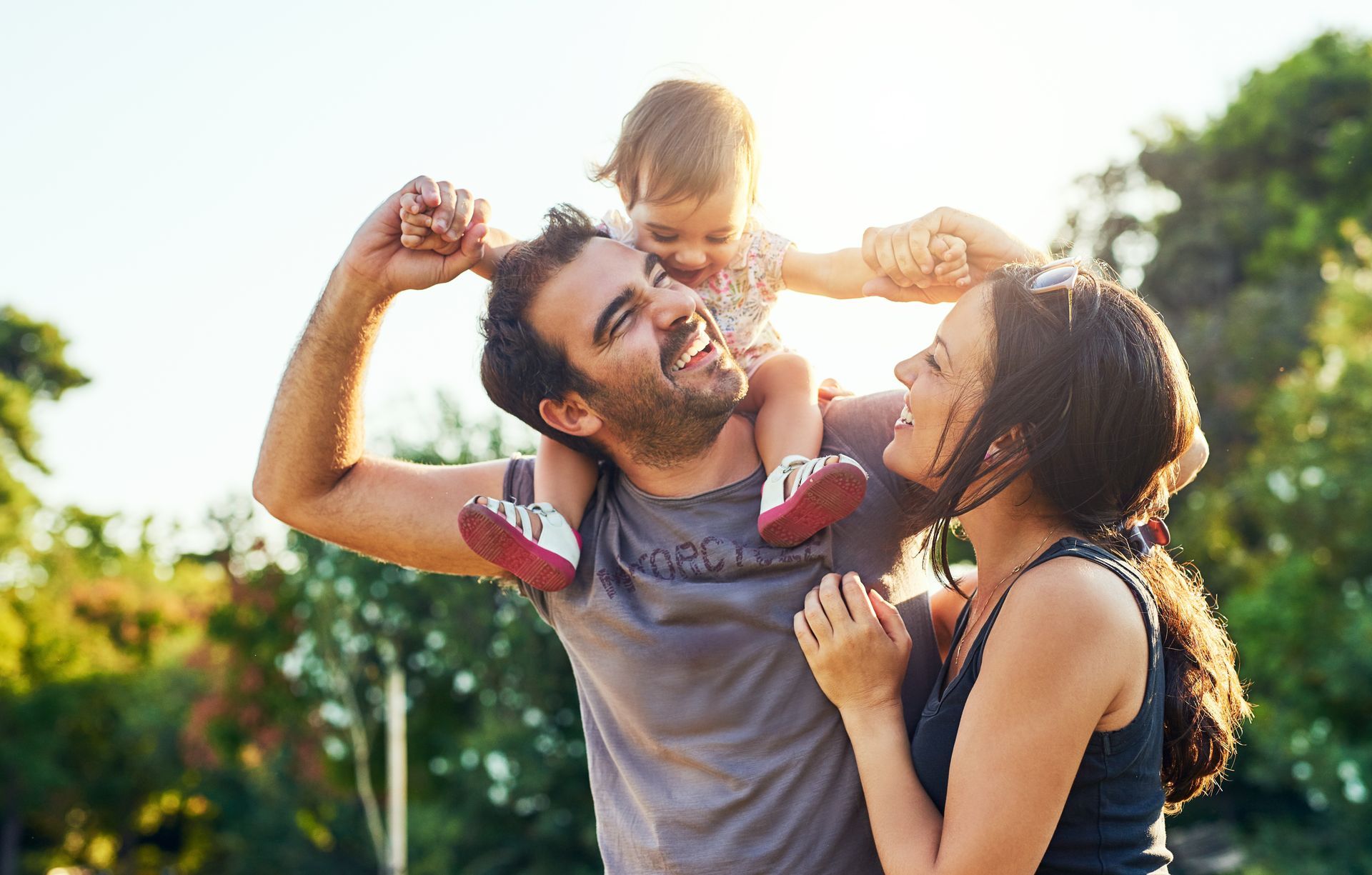Smiling family outdoors, parents lifting a child in the air on a sunny day.