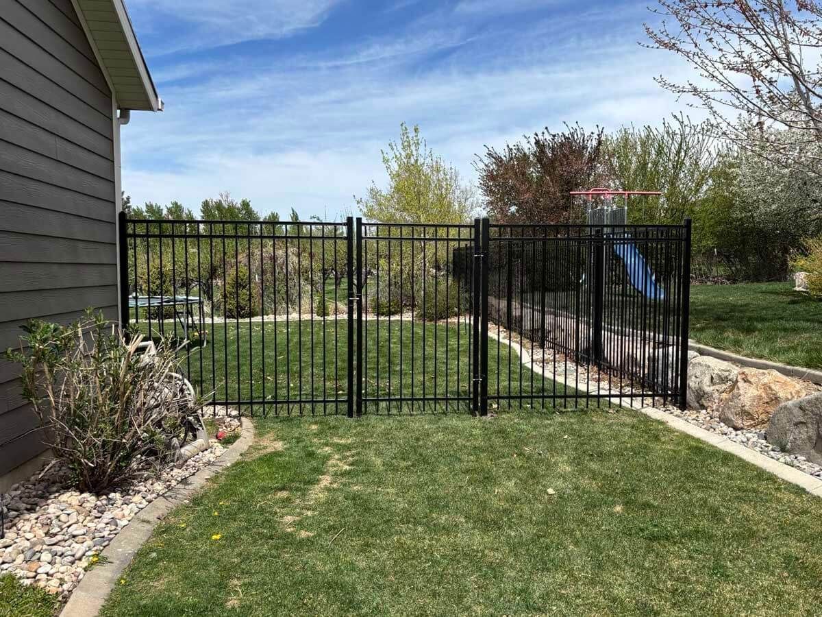 Black metal fence enclosing a grassy yard beside a house, with a stone border and trees behind it