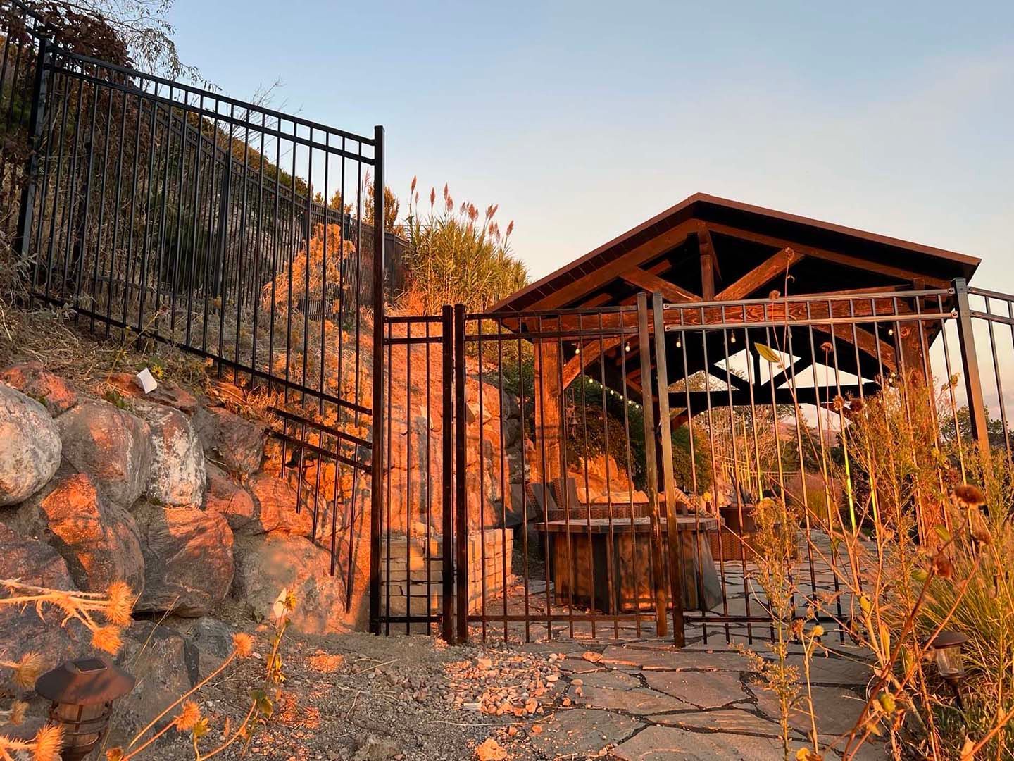 Stone wall and black gate beside a rustic pavilion with chairs, lit by warm sunset light.