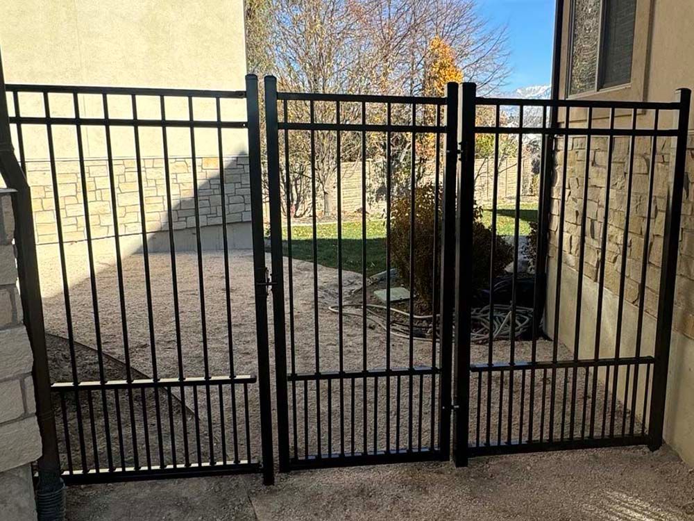 Black metal driveway gate between tan walls, with trees and lawn visible beyond.