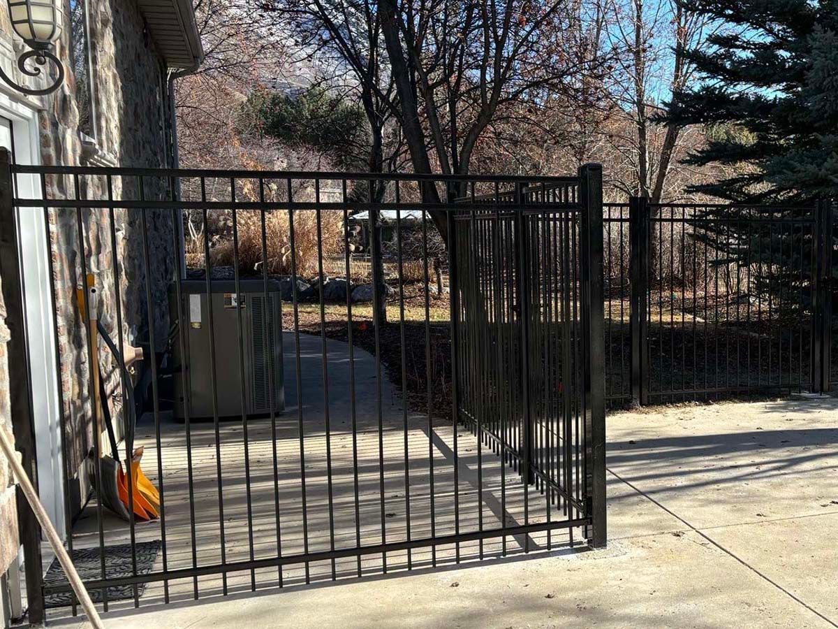 Black metal gate beside a concrete sidewalk in a residential yard, with trees and a brick building behind it