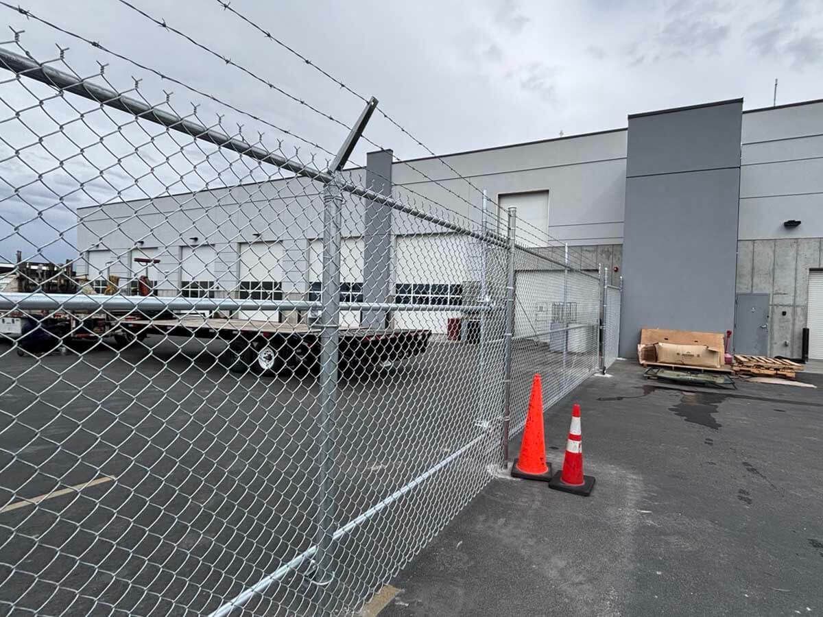 Chain-link fence beside industrial building, with orange cones and pallets in a paved lot.